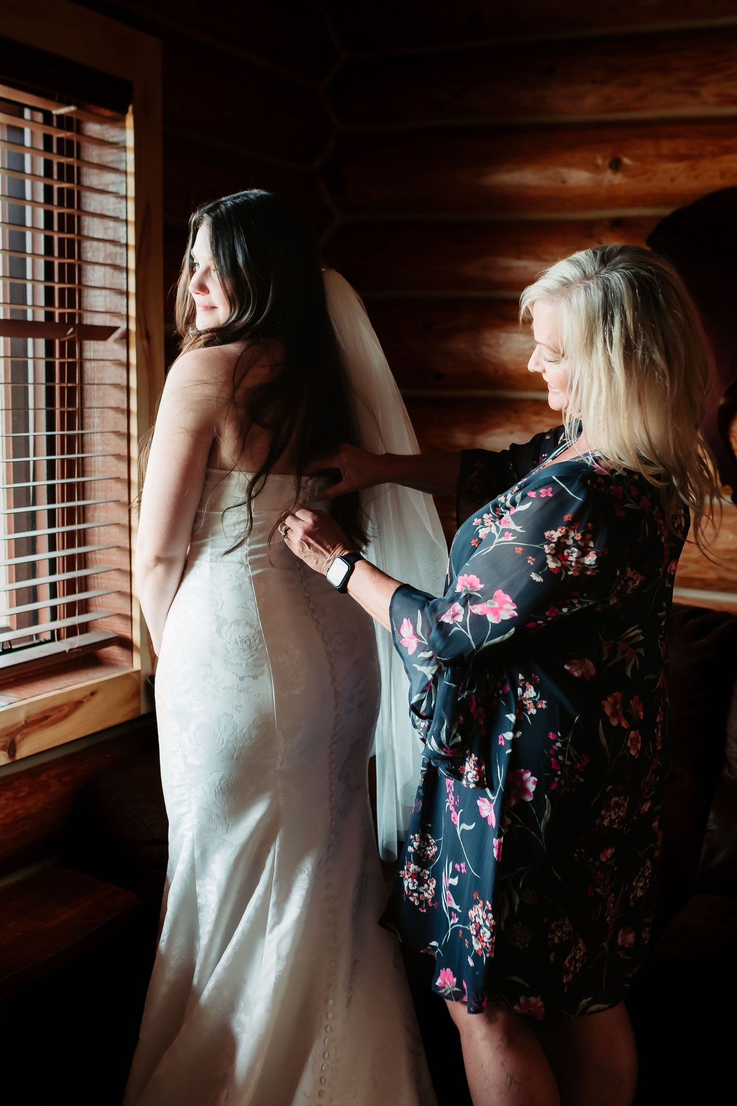 Woman in white wedding dress getting help with final adjustments in rustic log cabin setting near window.