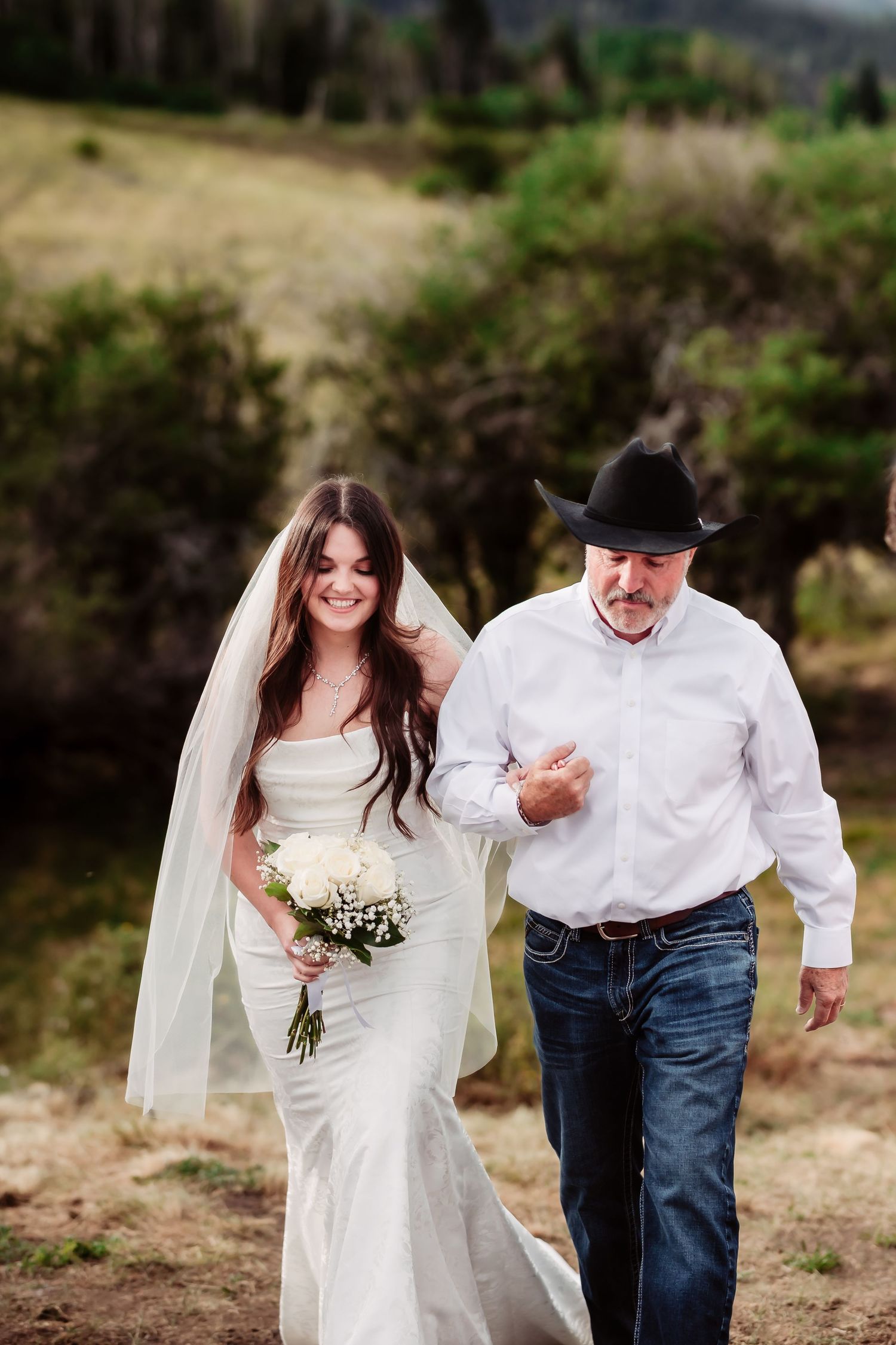 A bride in a white wedding dress holding a bouquet walks arm in arm with someone in a white shirt along a scenic path.