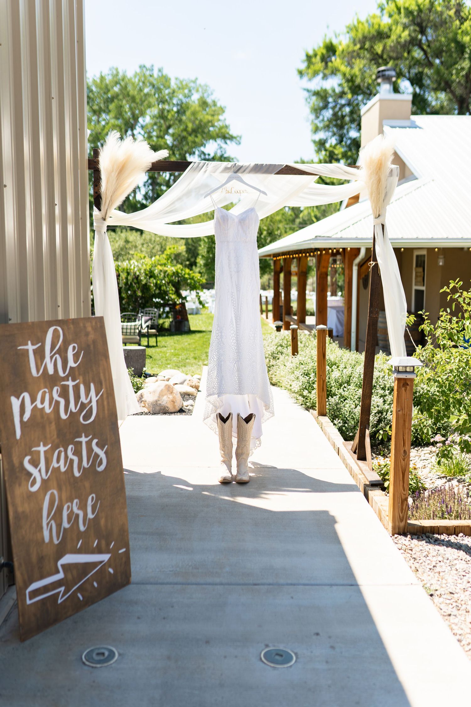 A white wedding dress hangs on display outdoors next to a wooden sign reading 'the party starts here'.