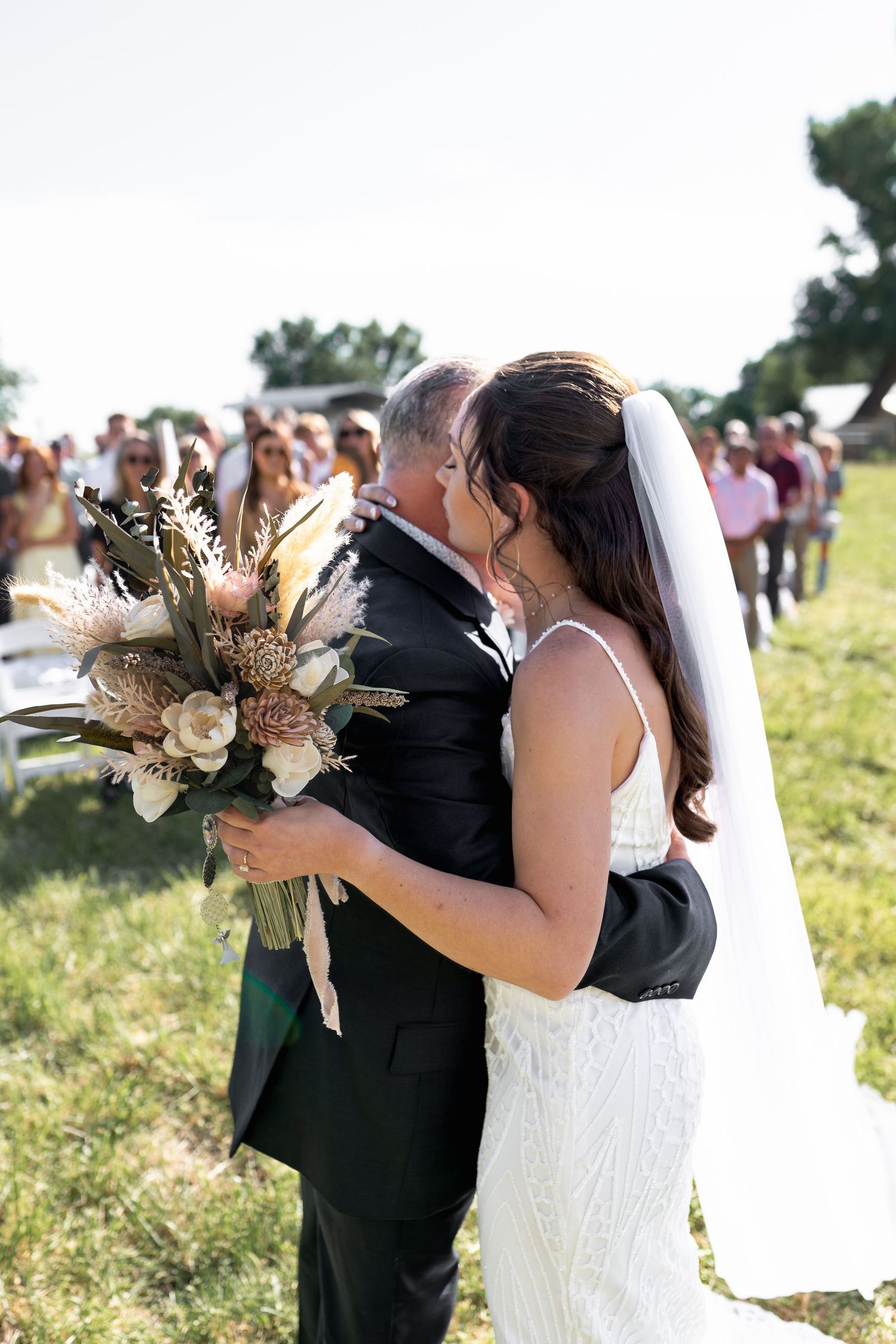 A touching moment during an outdoor wedding ceremony with the couple embracing and a dried floral bouquet.