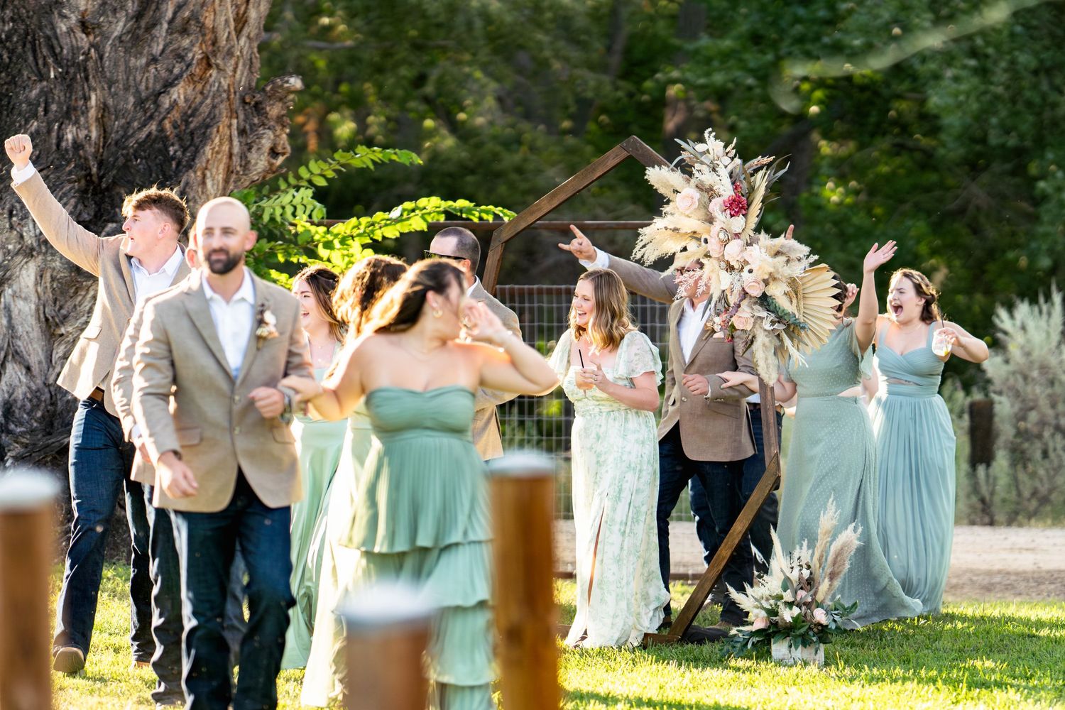 Wedding party celebrates joyfully on a sunny lawn with bridesmaids in mint green dresses raising their arms.