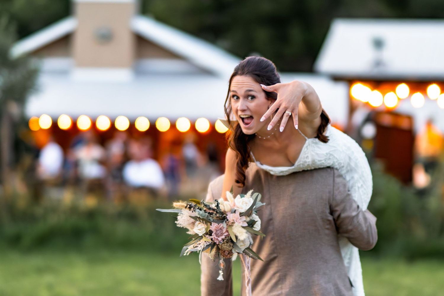 A person in a gray sweater shows off a ring while holding flowers at an outdoor evening event with twinkling lights.