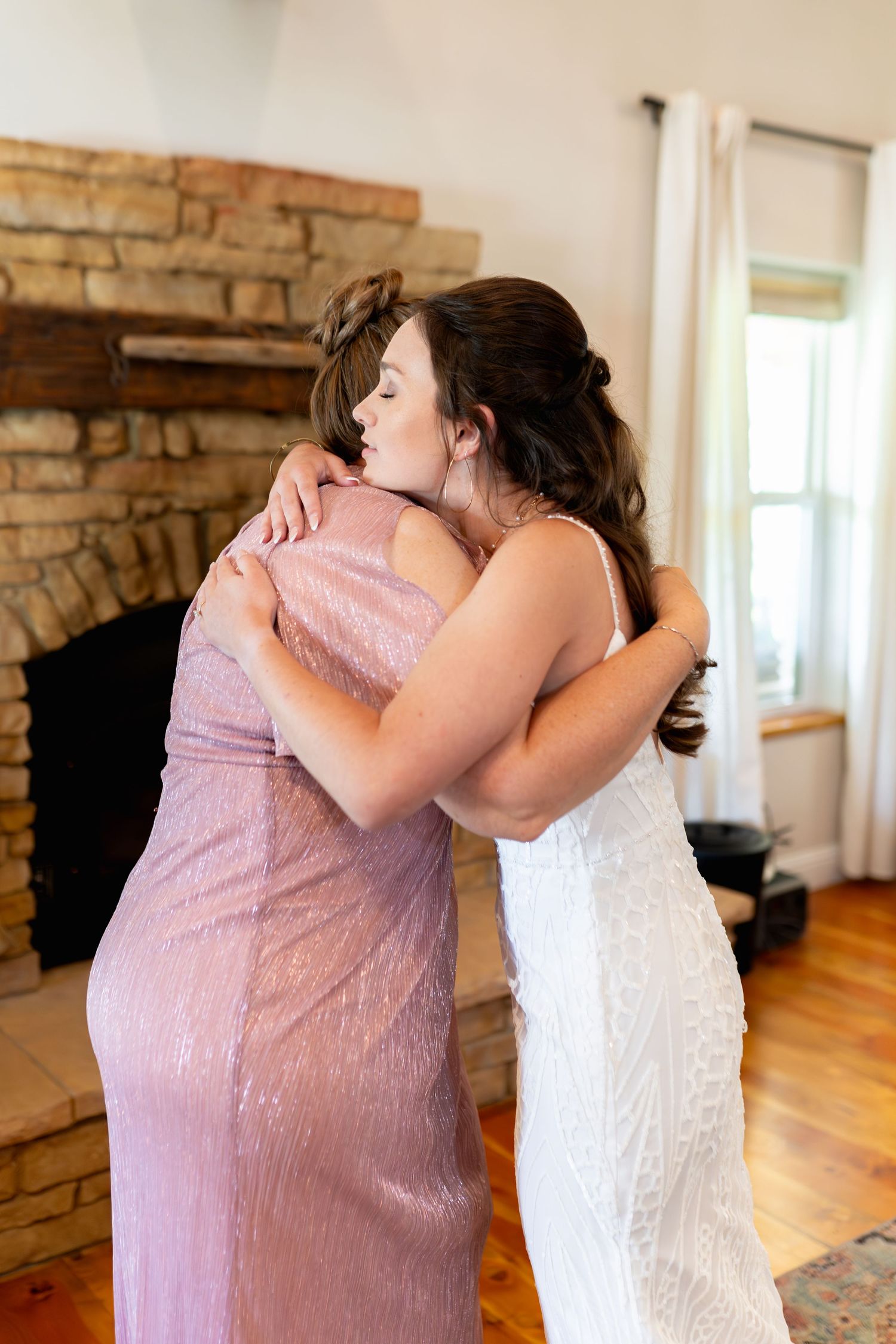 Two people embrace in an emotional moment, one wearing a pink dress and one in a white wedding gown beside a brick fireplace.