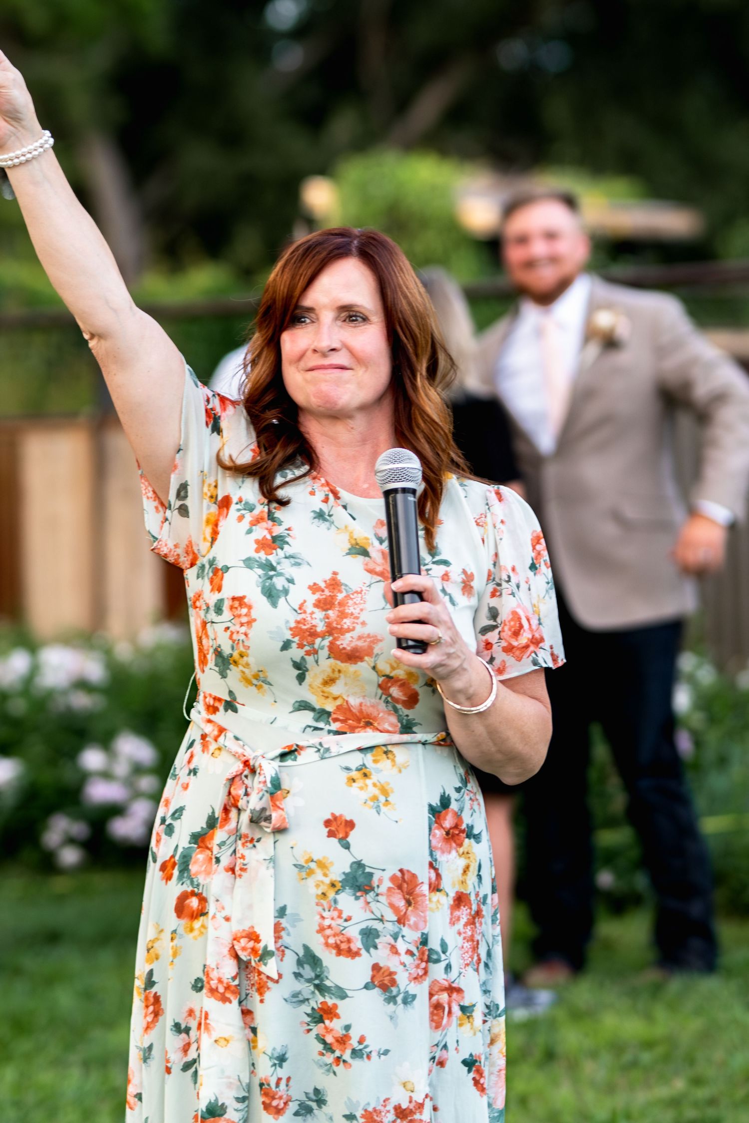 A speaker in a floral dress enthusiastically addresses the wedding crowd with microphone in hand.