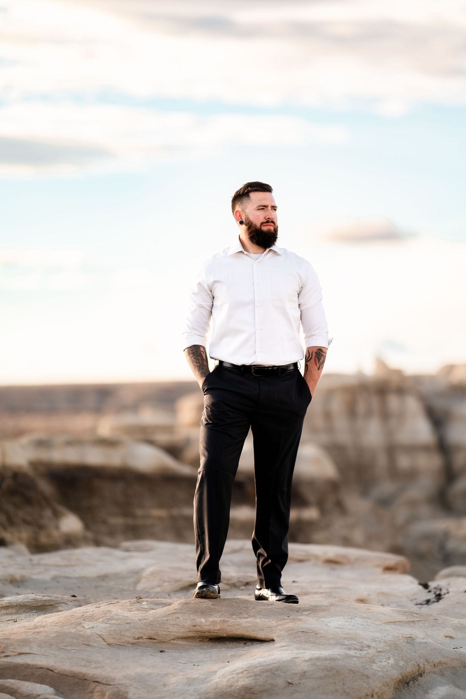 A well-dressed man in a white shirt and dark pants stands confidently on rocky terrain against a cloudy sky.