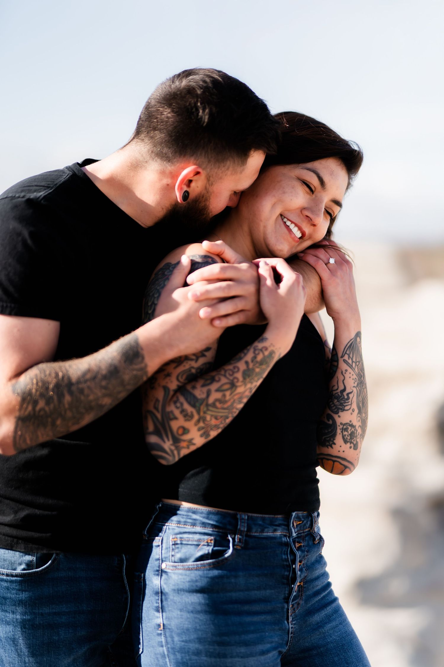 A couple shares a playful moment together, showcasing tattooed arms and casual attire.