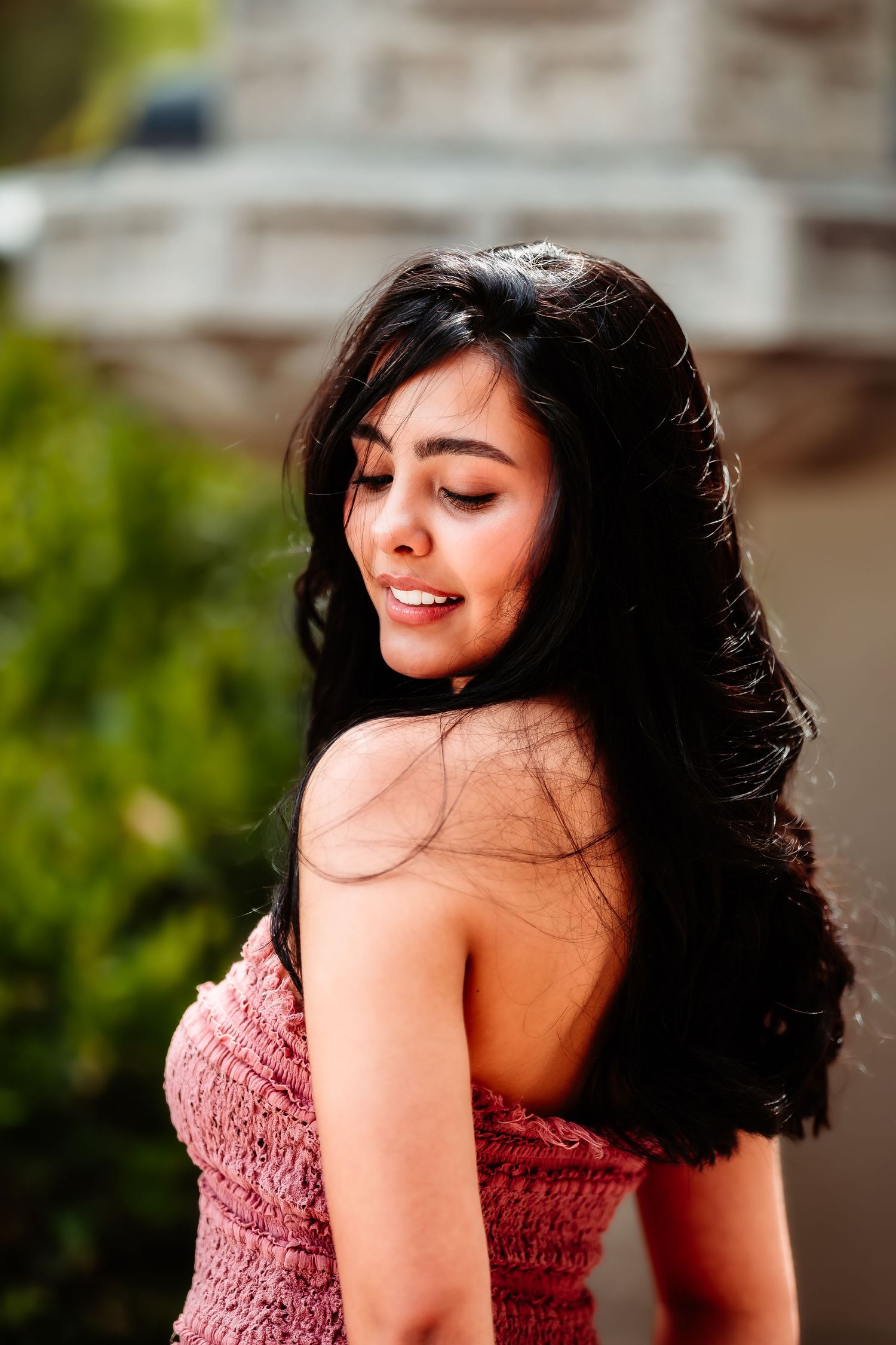 Woman in pink strapless dress smiling while looking down with long dark hair flowing in natural light.