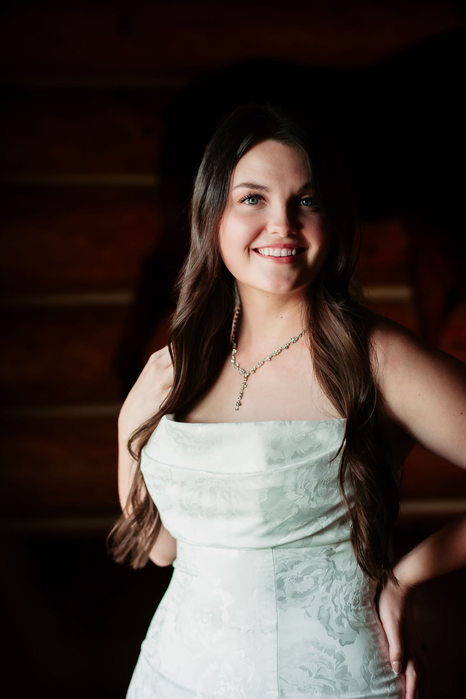Woman in elegant white lace dress poses in dramatic lighting against dark wooden backdrop.