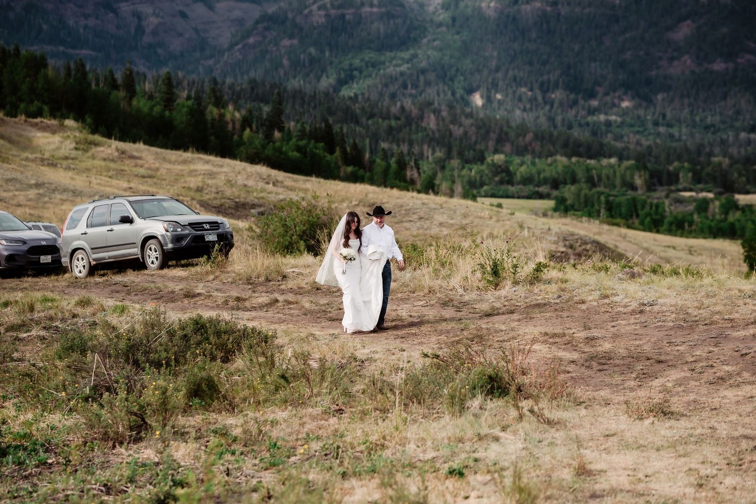 A couple in wedding attire walks along a mountain trail with parked cars in the distance.