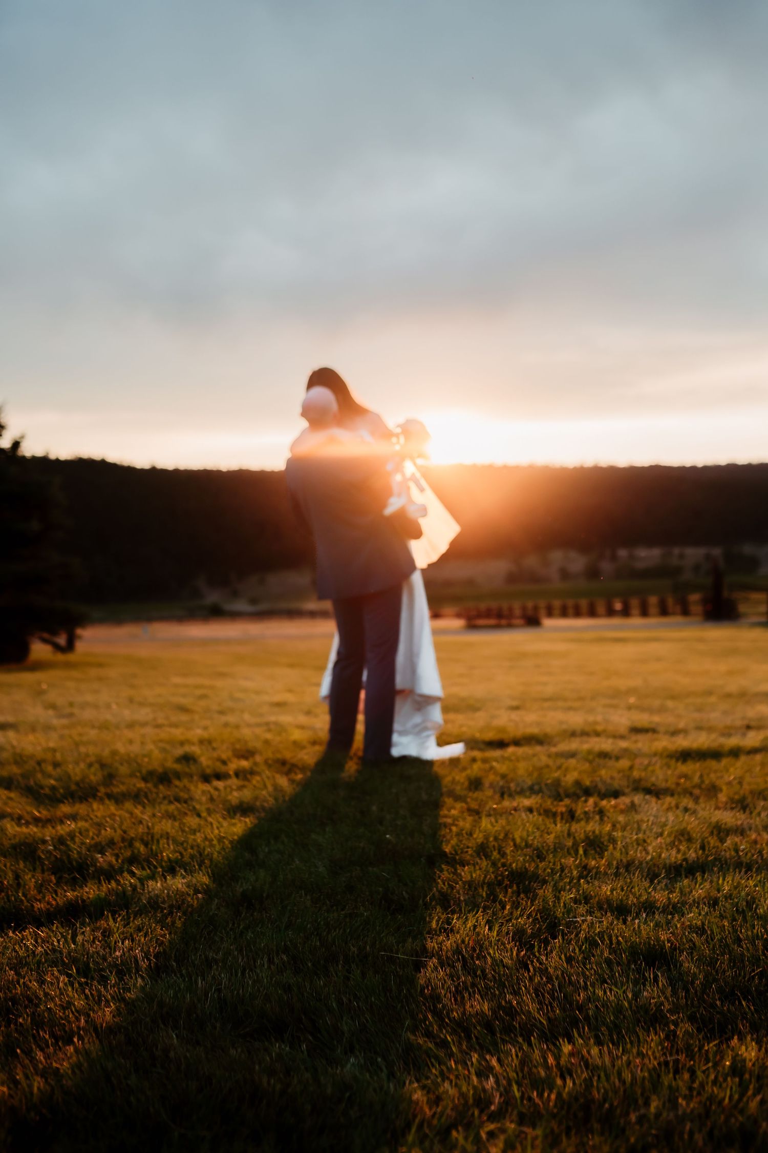 Silhouetted wedding couple embraces against golden sunset in mountain meadow.