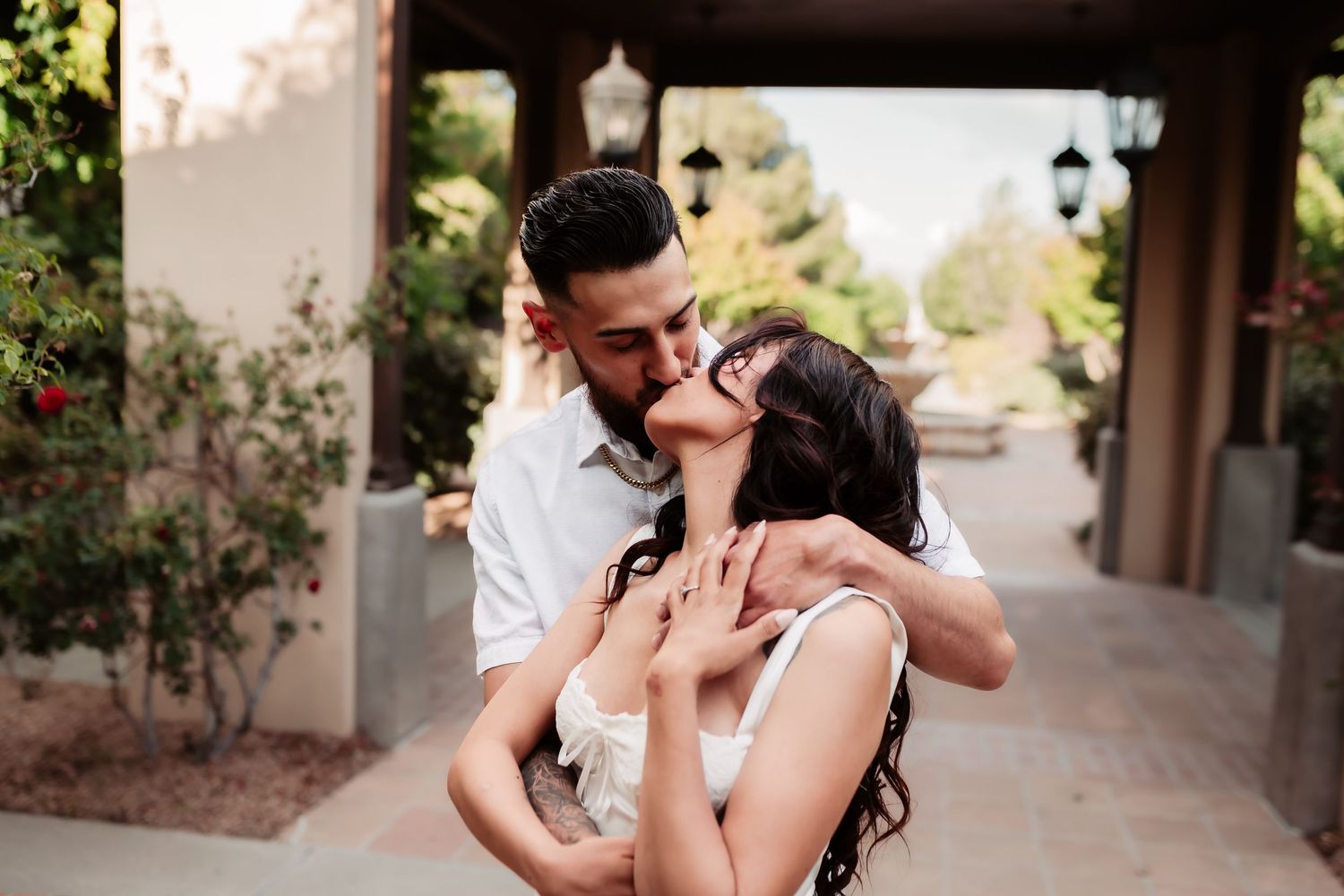 A couple shares an intimate embrace under an outdoor pergola surrounded by soft natural lighting.