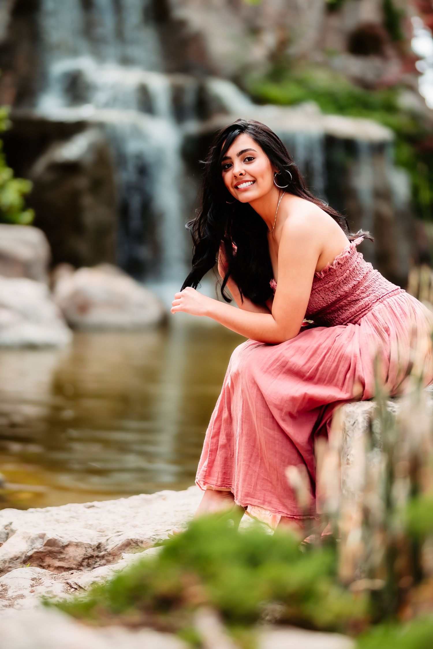 A person in a pink flowy dress sits gracefully on rocks near a serene waterfall in a natural outdoor setting.