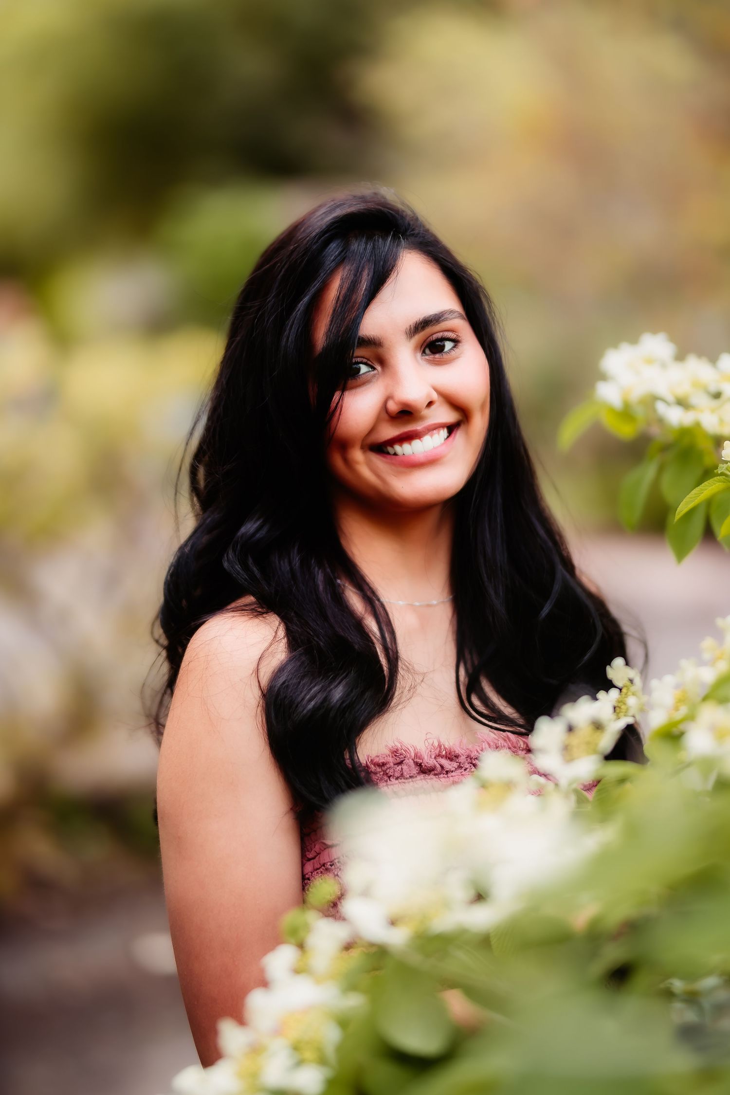 A person with long dark hair poses among white flowers wearing a pink top in a garden setting.