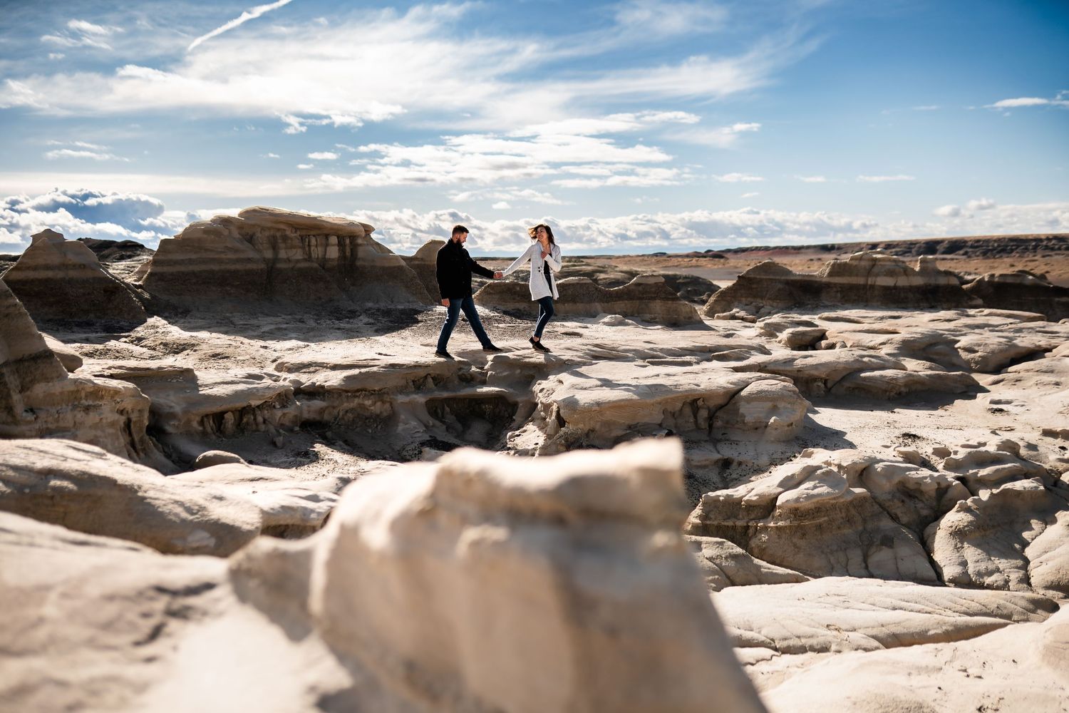 Two silhouettes walk across dramatic desert rock formations under a cloudy blue sky.