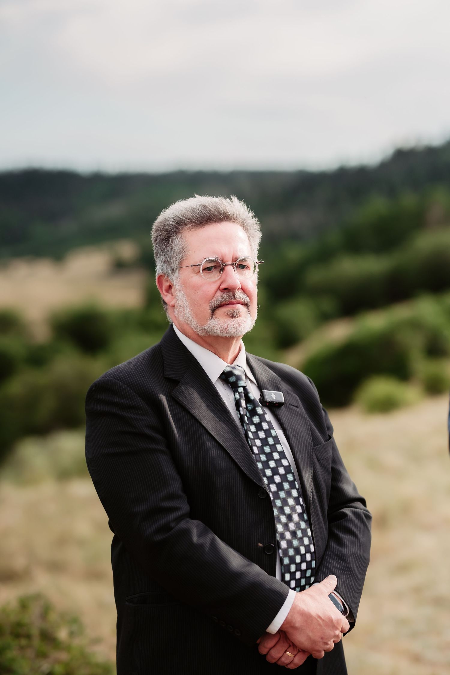 An officiant in a black suit with polka dot tie stands outdoors with rolling hills in the background.