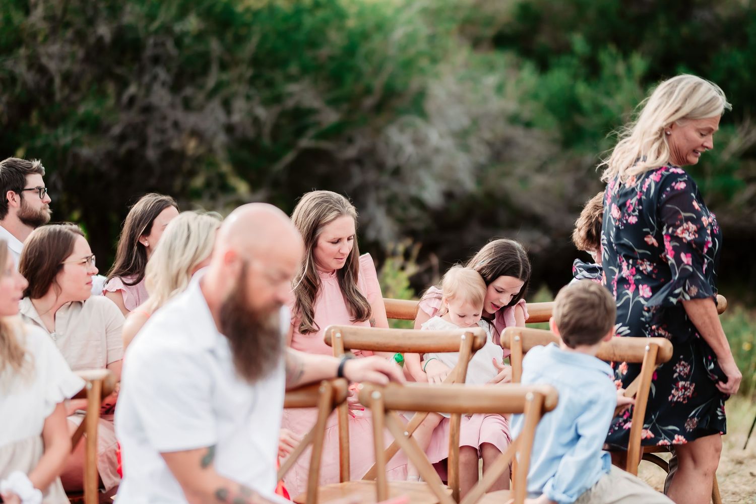 Wedding guests sitting in wooden farmhouse chairs at an outdoor ceremony surrounded by greenery.
