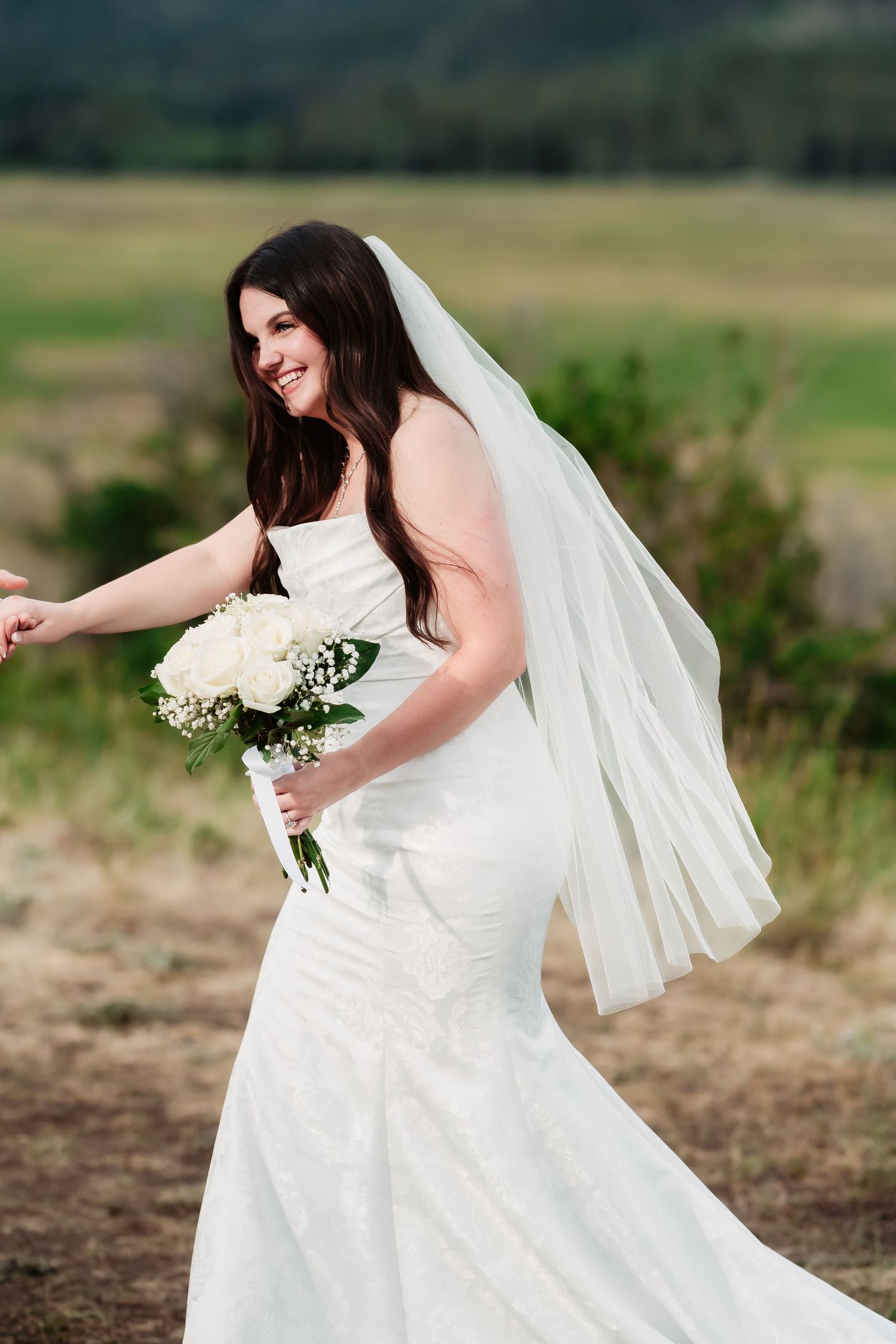 A joyful bride in a fitted white gown holds her bouquet against a mountain backdrop.