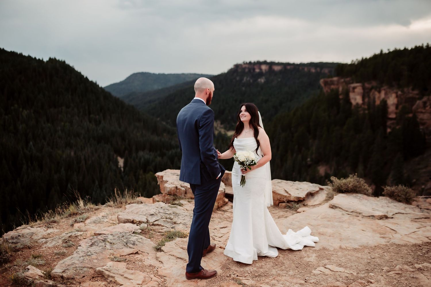 A couple shares a moment on a clifftop overlooking a dramatic mountain valley.