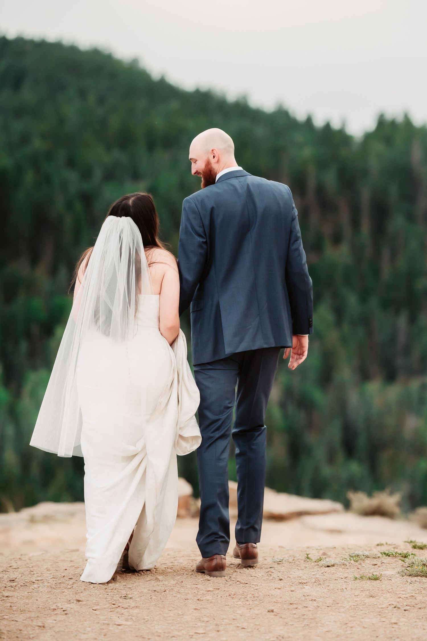 A bride and groom walking away together on a mountain trail with evergreen forest in the background.