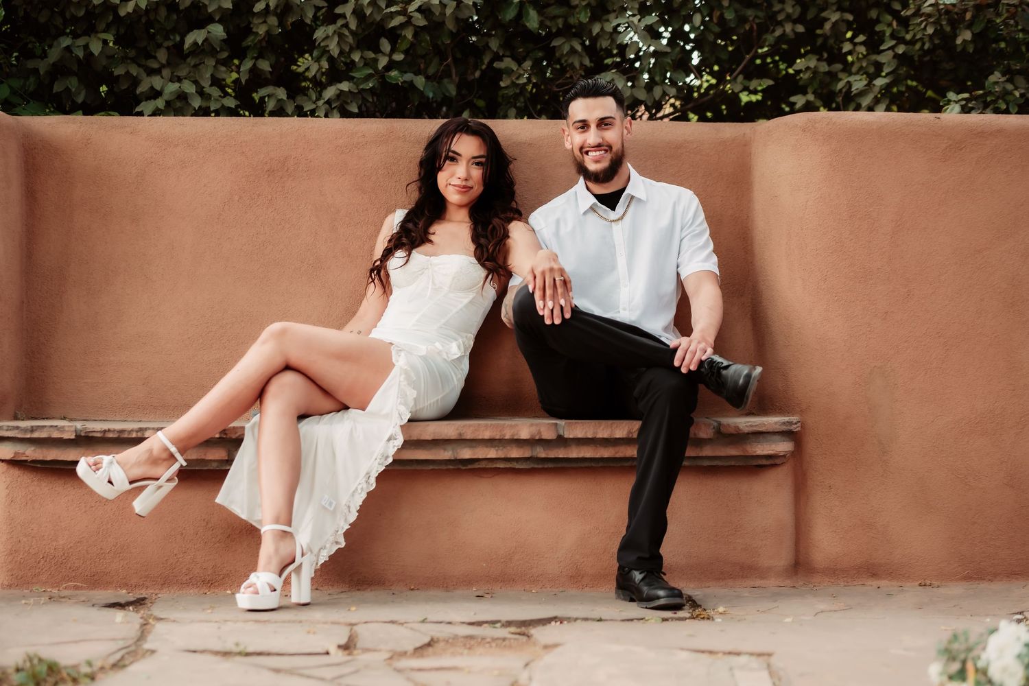 Stylish couple in white lounging on terracotta bench during engagement session.