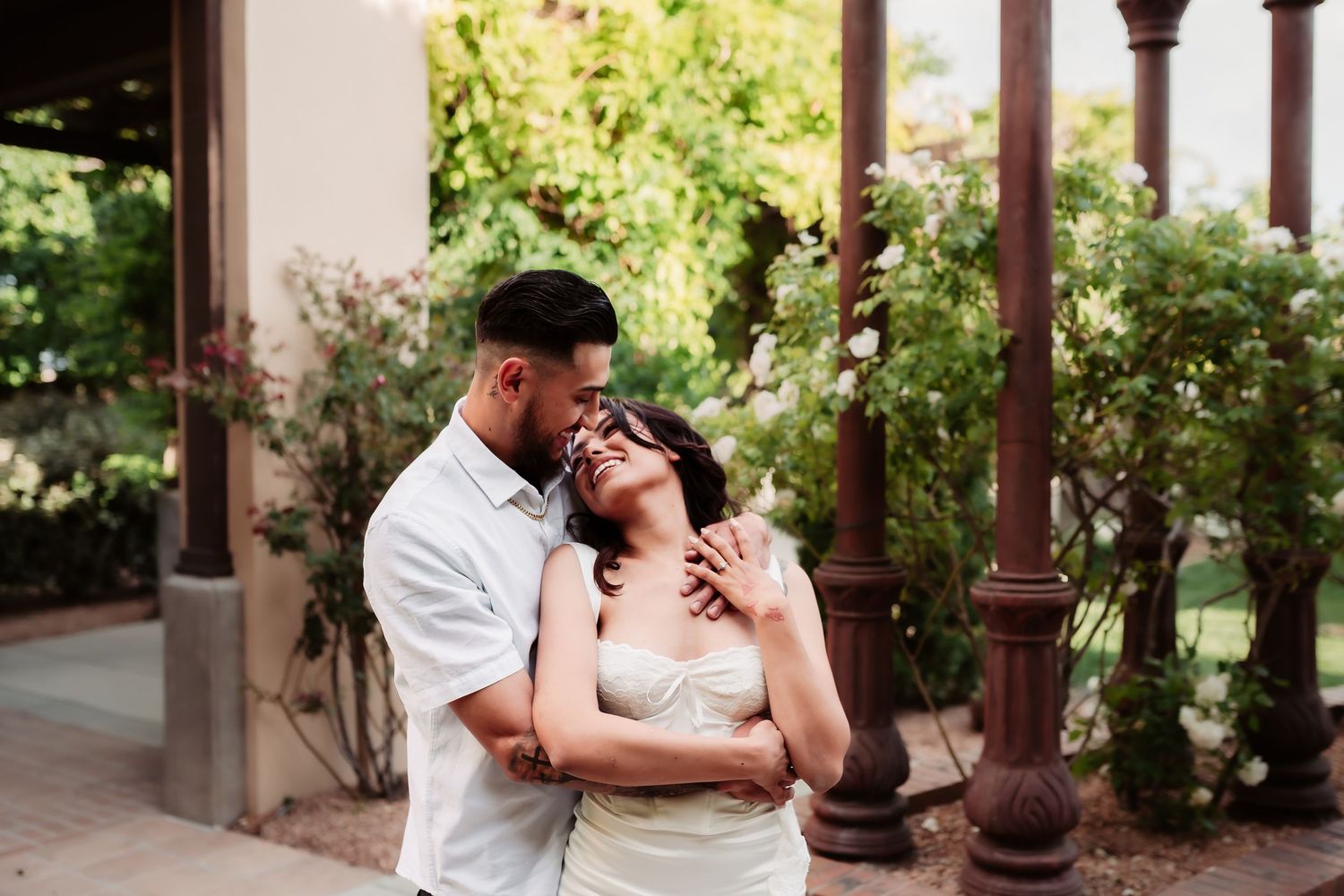 A couple embraces intimately under a covered walkway surrounded by lush garden foliage.