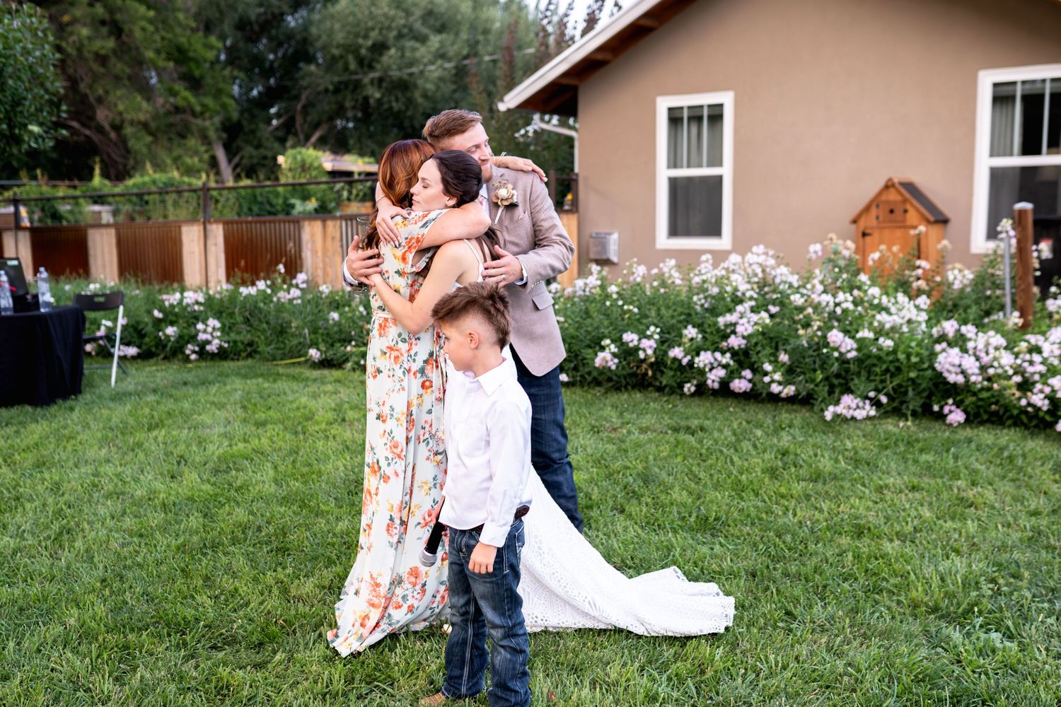 Three people share a sweet embrace on a grassy lawn with white flowers and a pink house in the background.