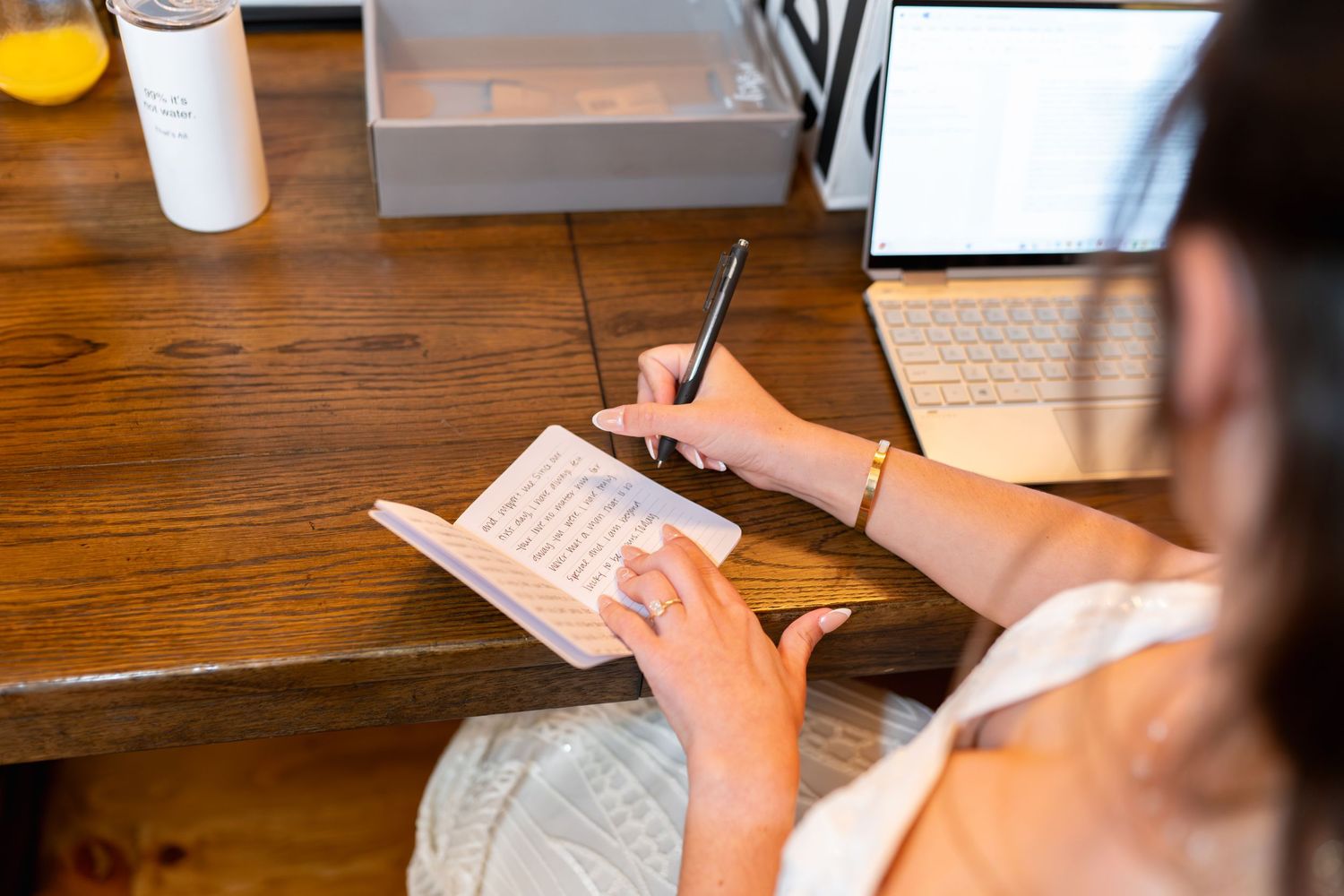 Person writing with pen on paper at wooden desk with coffee cup and laptop in background.