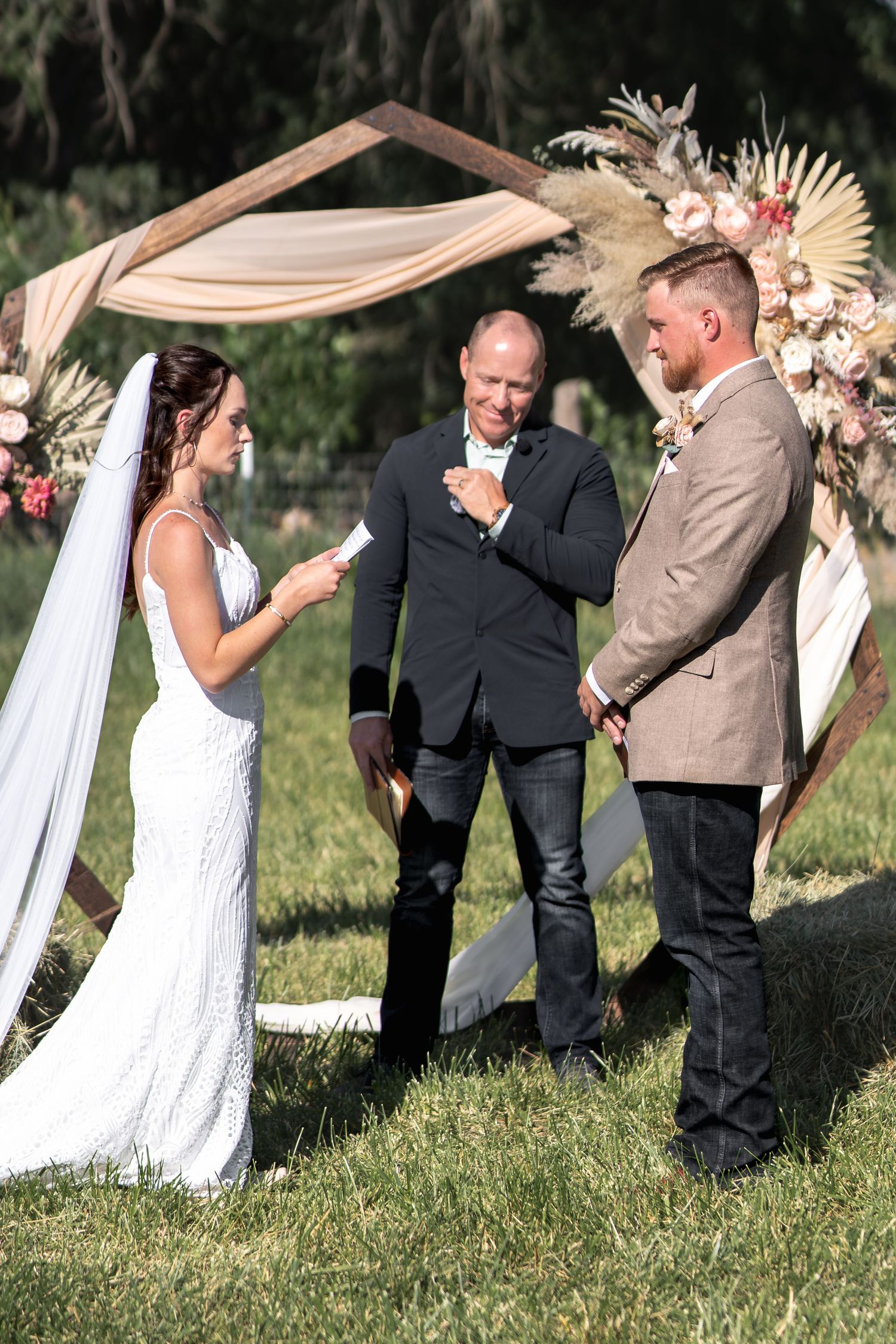 An outdoor wedding ceremony under a decorated arch with dried flowers and fabric draping.