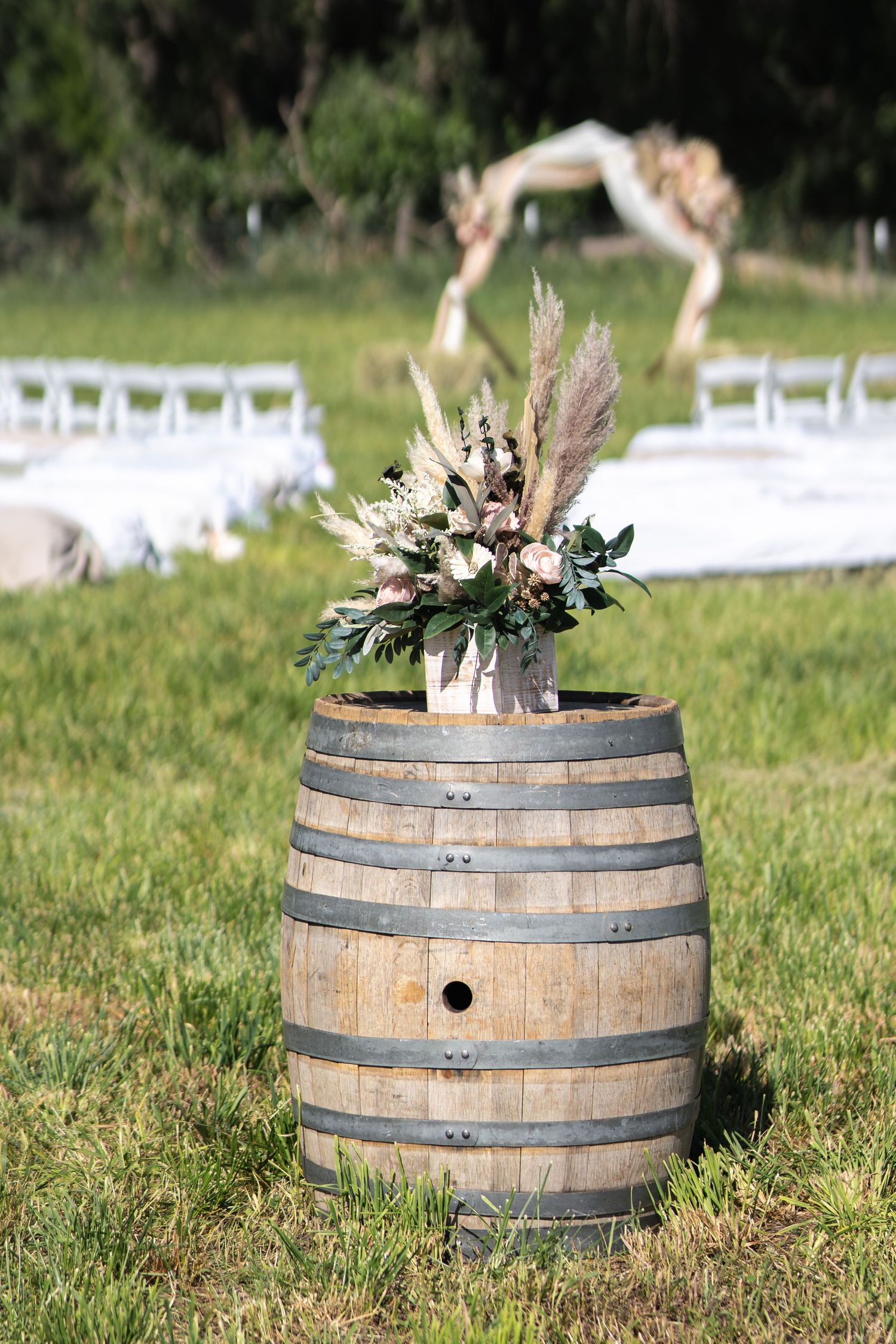 Rustic wine barrel decorated with dried floral arrangement at outdoor wedding ceremony with white chairs.