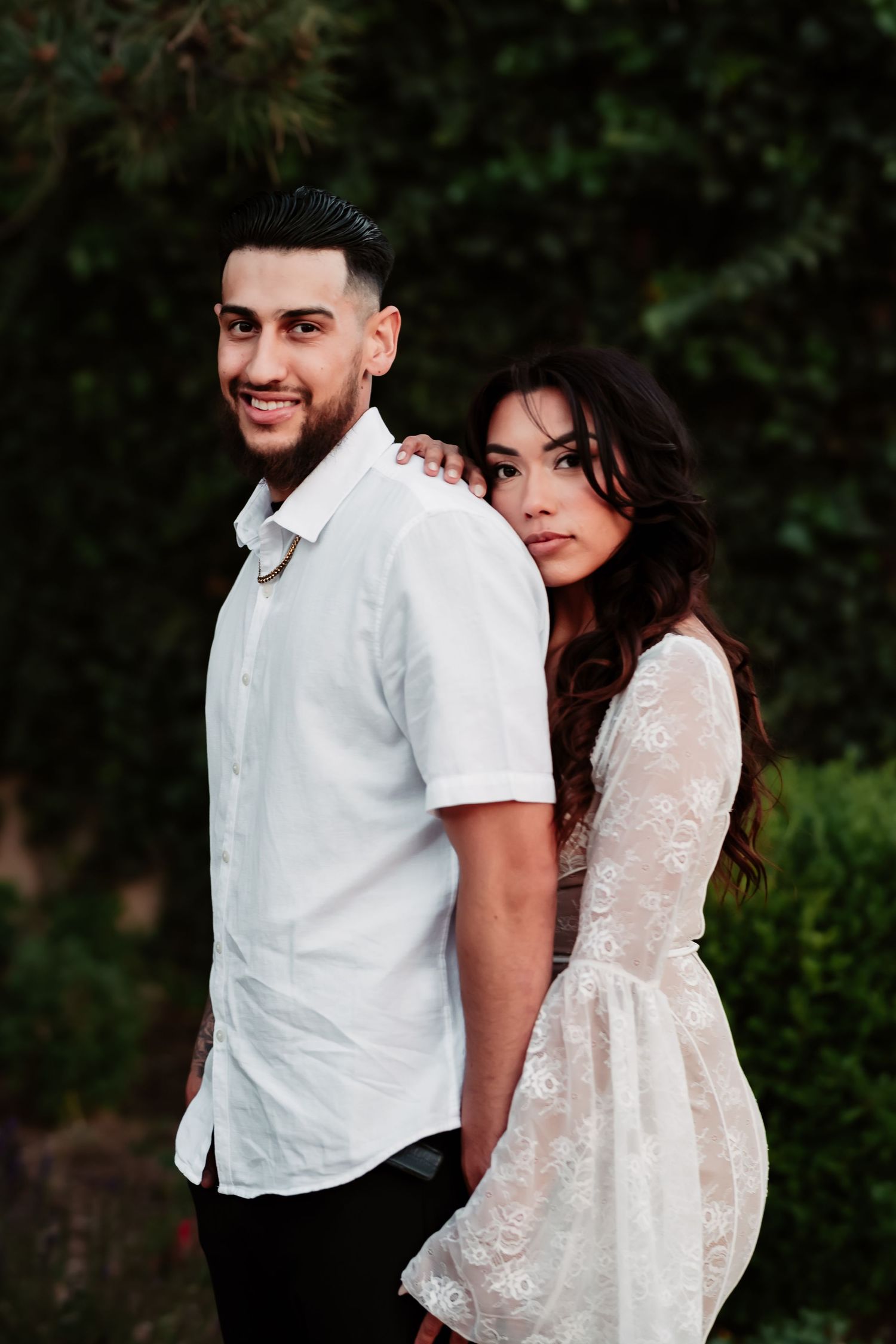 A stylish couple poses outdoors in coordinated white outfits against lush greenery.