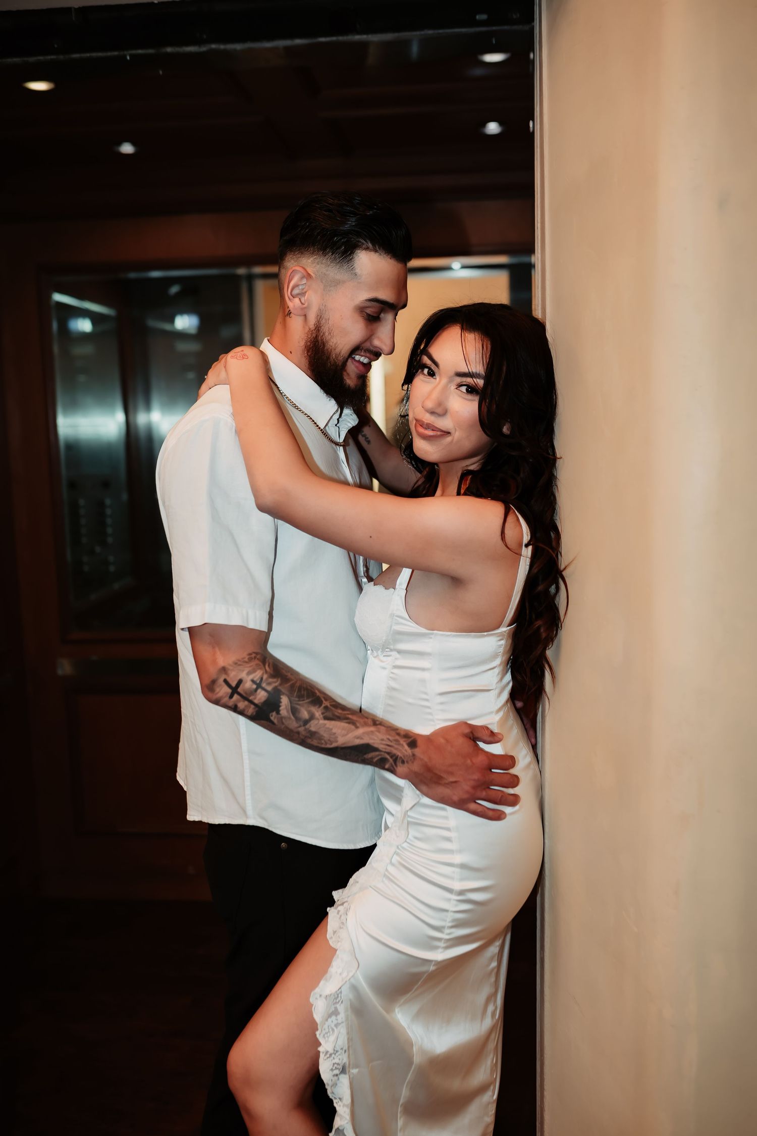 A couple in formal attire share an intimate moment in an indoor hallway setting.