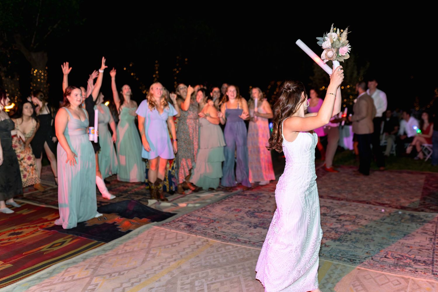 Bride tosses bouquet during nighttime reception as wedding guests gather on decorated outdoor carpet.