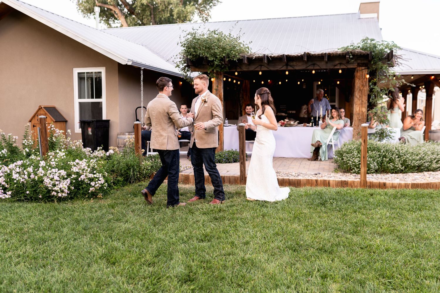 Casual outdoor wedding gathering with guests mingling on a lawn near a rustic building.