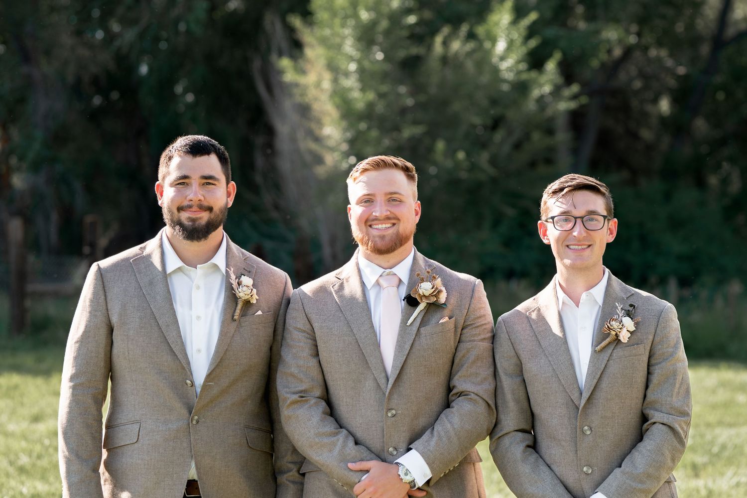 Three groomsmen wearing light gray suits and boutonnieres pose together outdoors during a wedding.