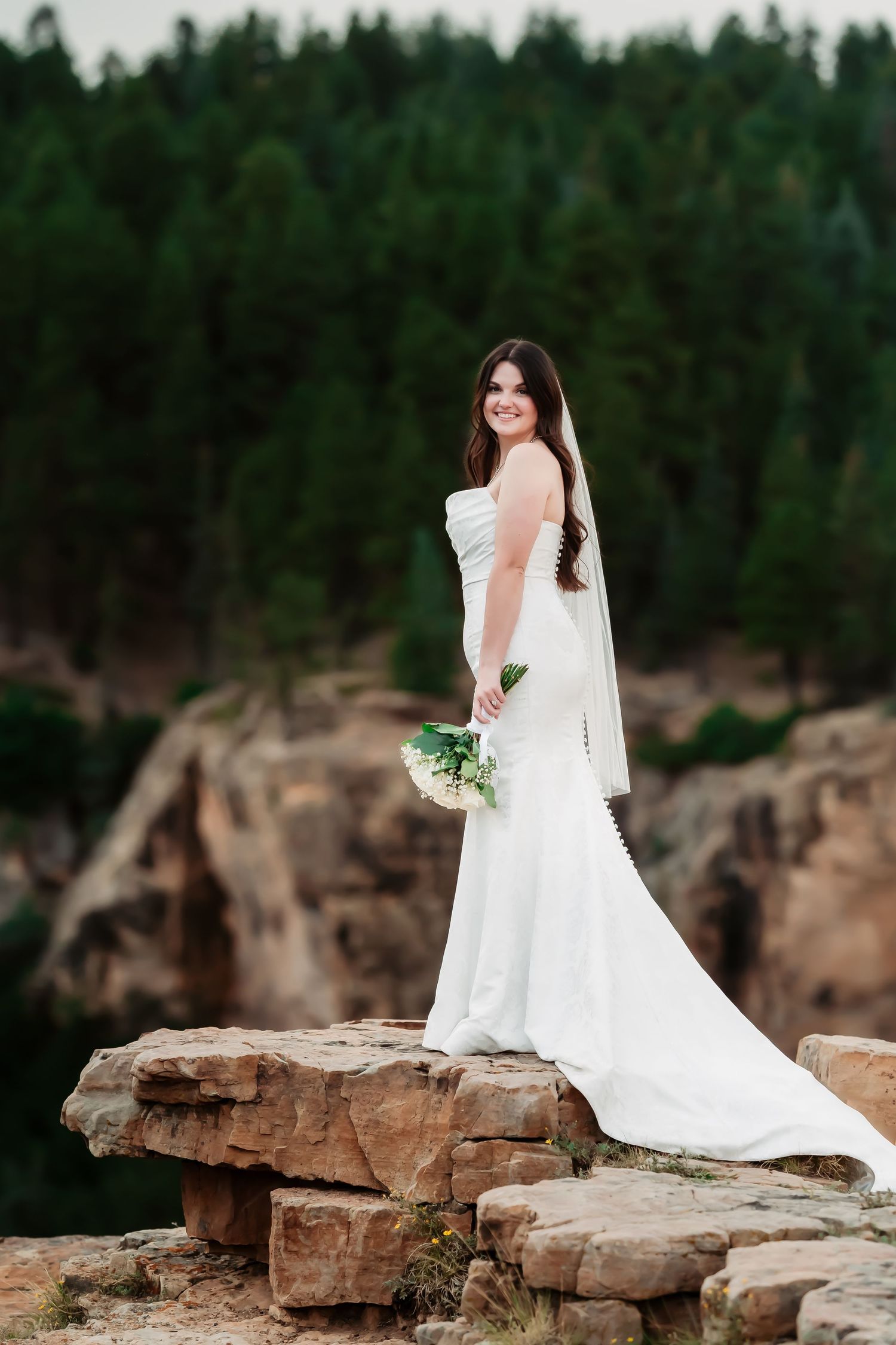 Bride in flowing white dress poses elegantly on rocky mountain outcrop.