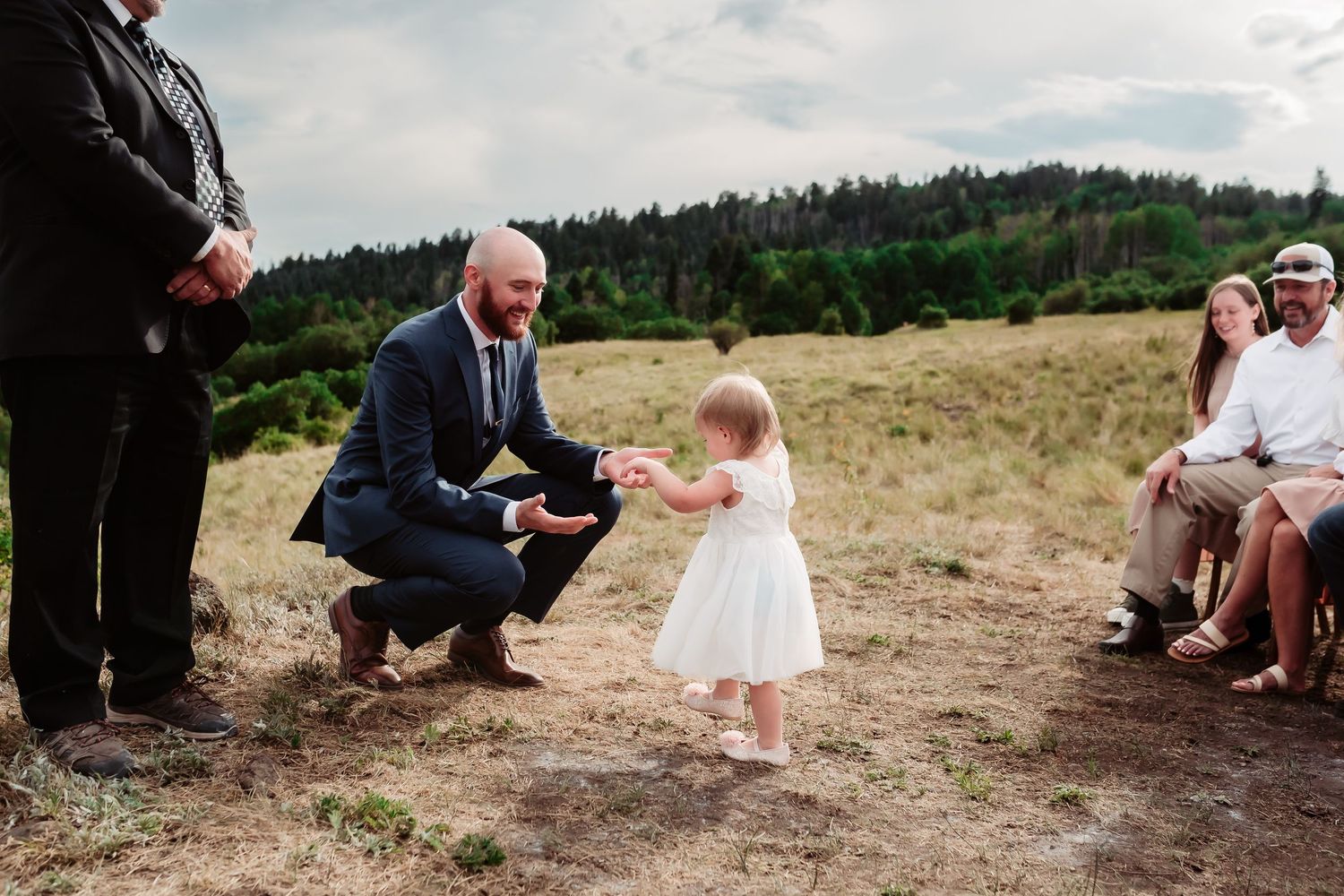 A touching moment during a wedding ceremony with guests seated on wooden chairs in a mountain setting.