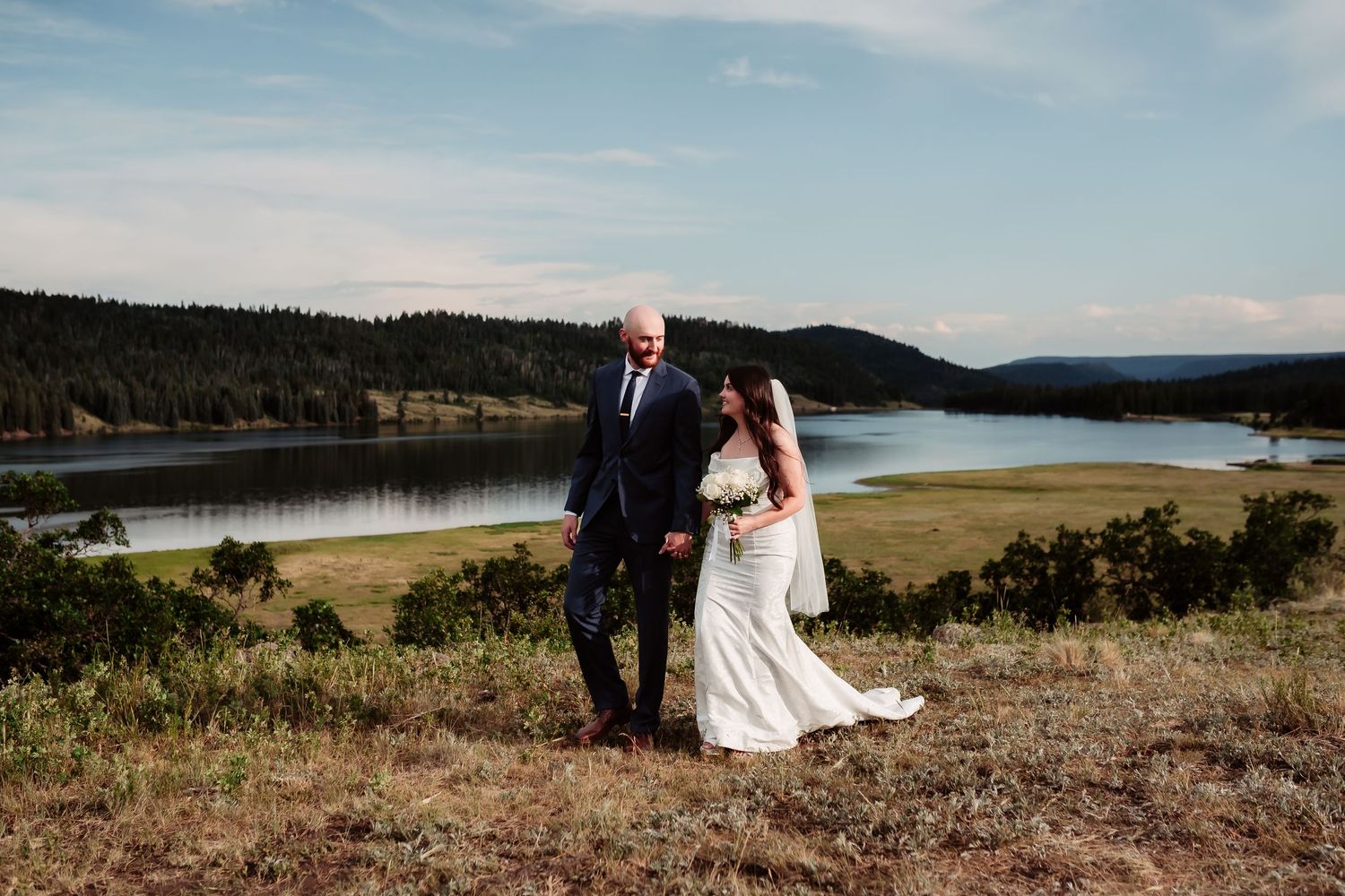 A wedding couple stands together on a hill with a beautiful lake and mountain vista behind them.