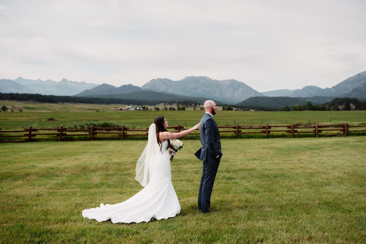 A couple stands together in a scenic mountain meadow on their wedding day with wood fences and rolling hills.