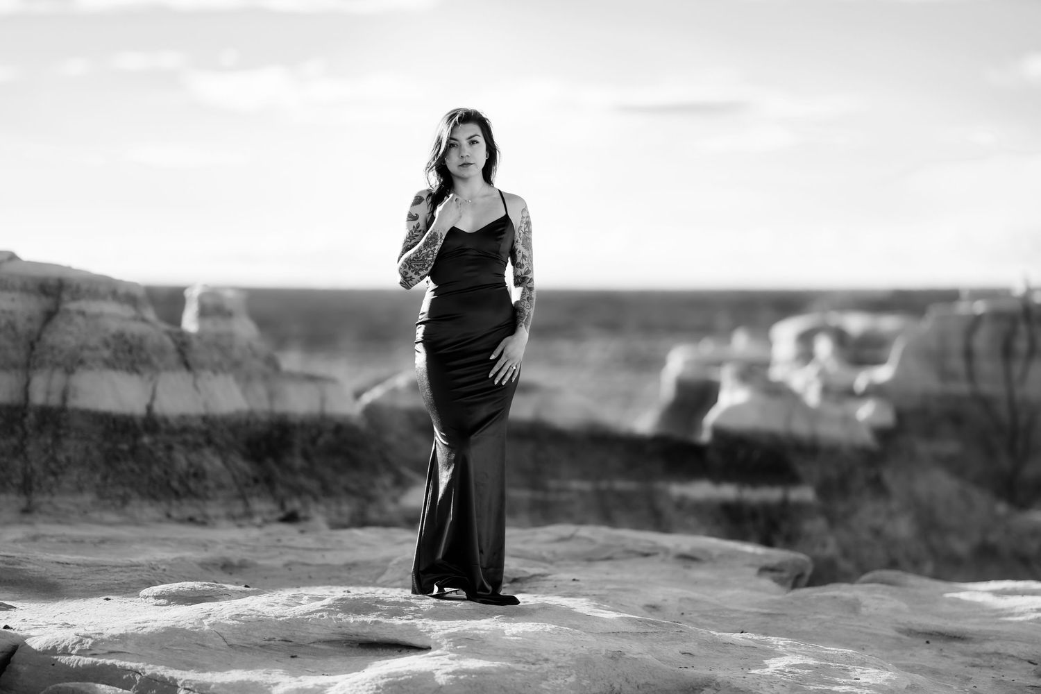 A figure in a flowing black dress stands alone on a rocky overlook in black and white.