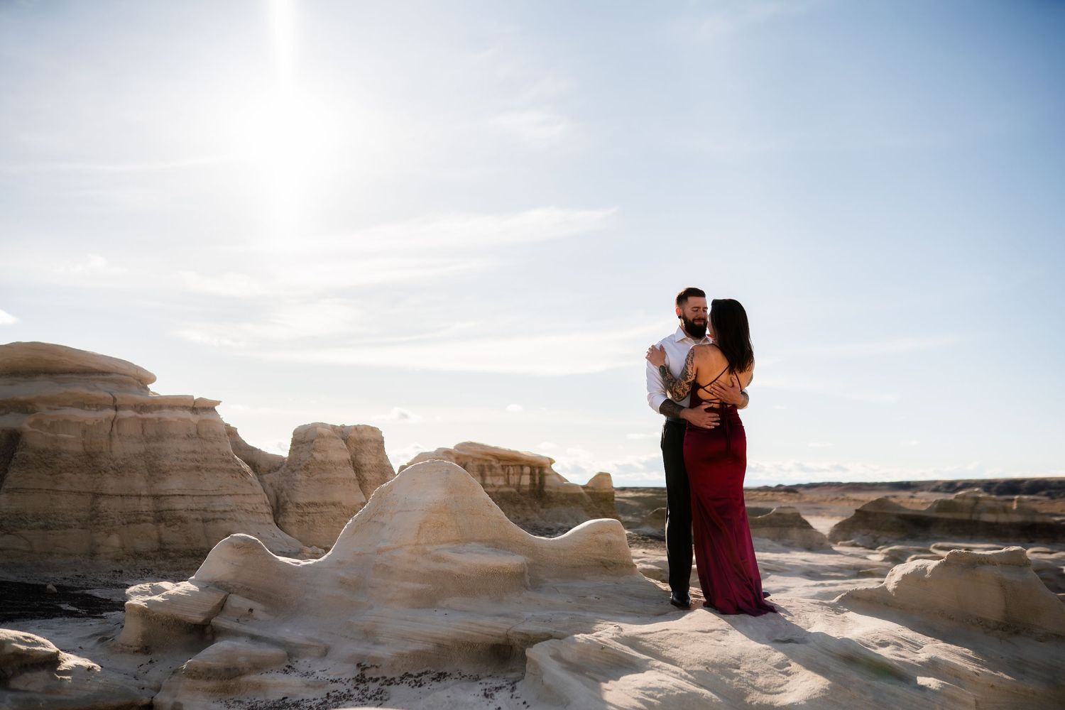 A couple in a burgundy dress embrace against smooth eroded rock formations under a bright sky.