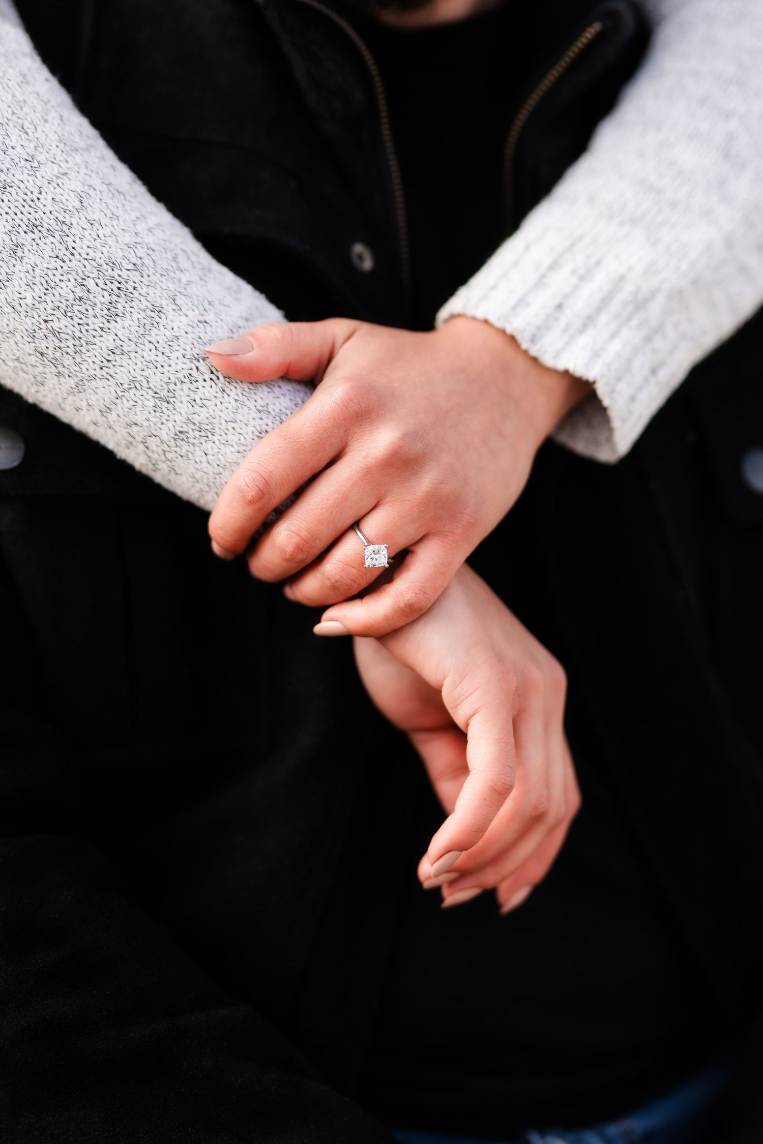 A close-up shot of hands wearing a white knit sweater and showcasing an engagement ring.