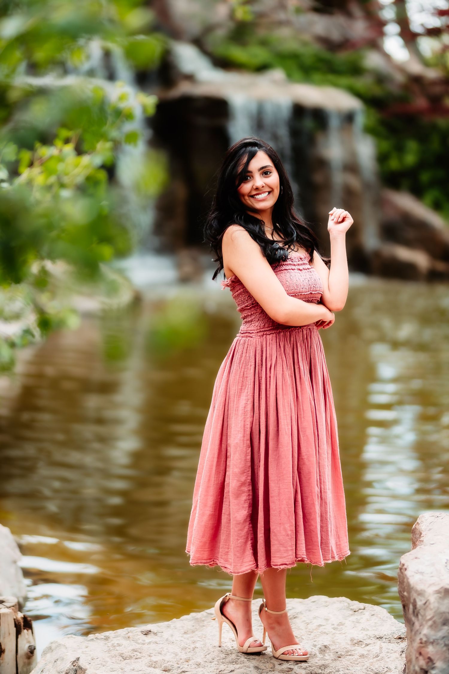 A person in a pink midi dress poses elegantly by a waterfall pond.