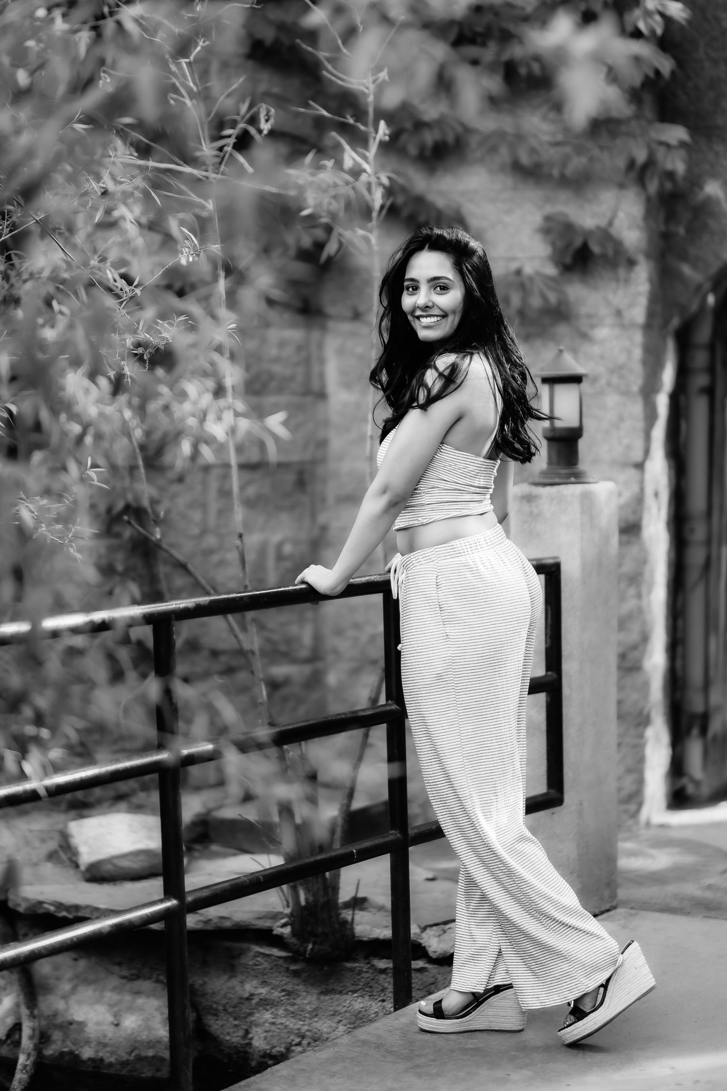 A black and white portrait of someone in casual wear smiling while standing by a railing outdoors.