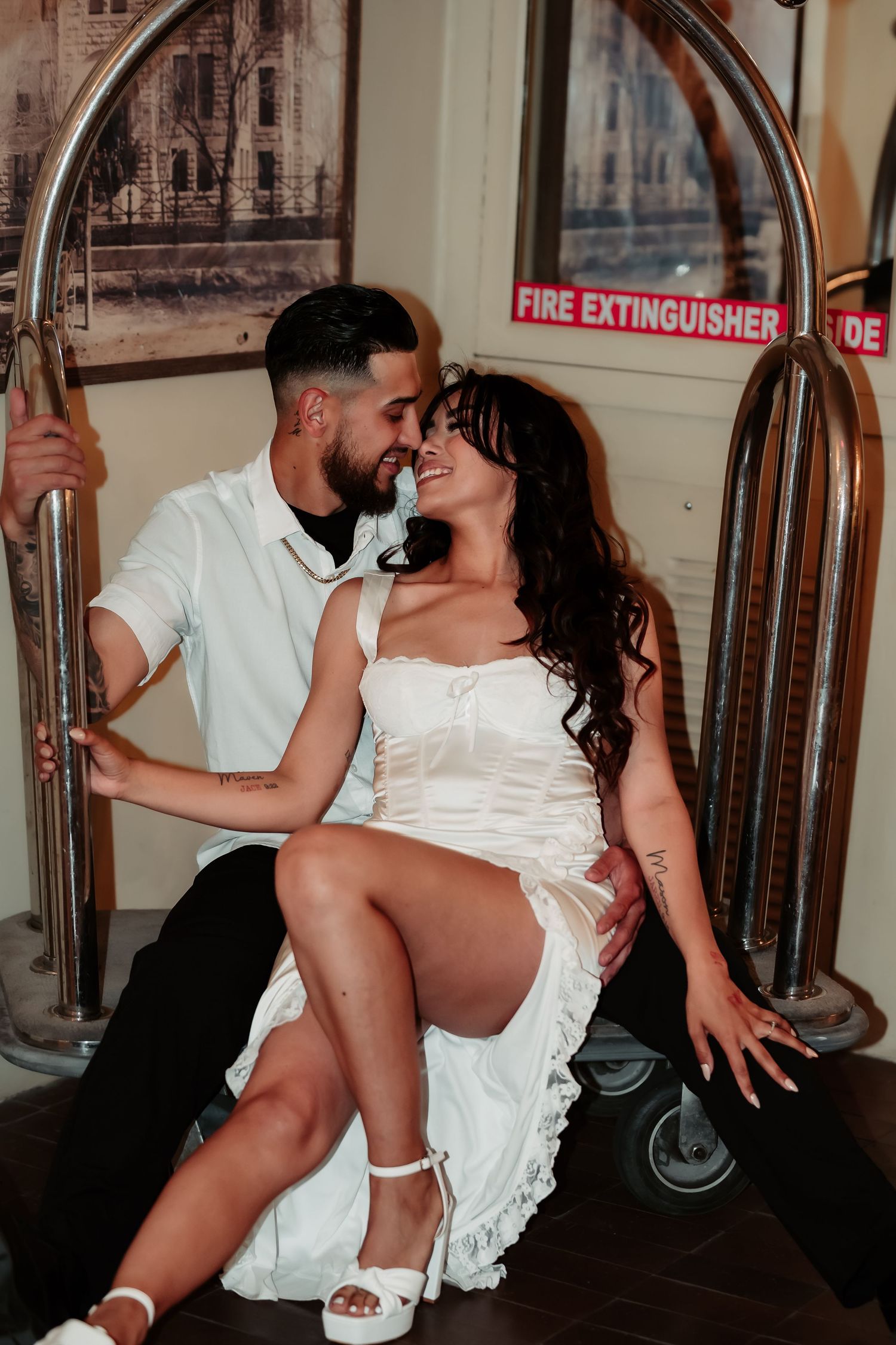 Romantic couple sharing an intimate moment on luggage cart with a fire extinguisher sign visible in background.