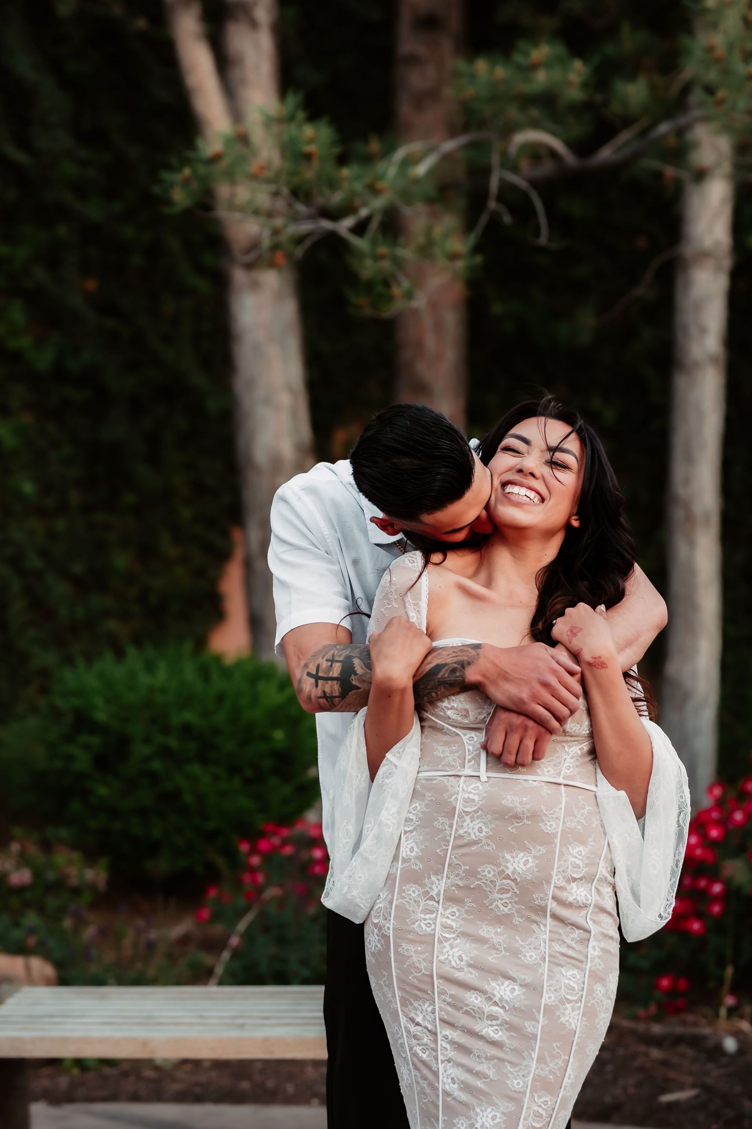 Romantic couple laughs joyfully in a garden setting, wearing formal attire as they embrace.