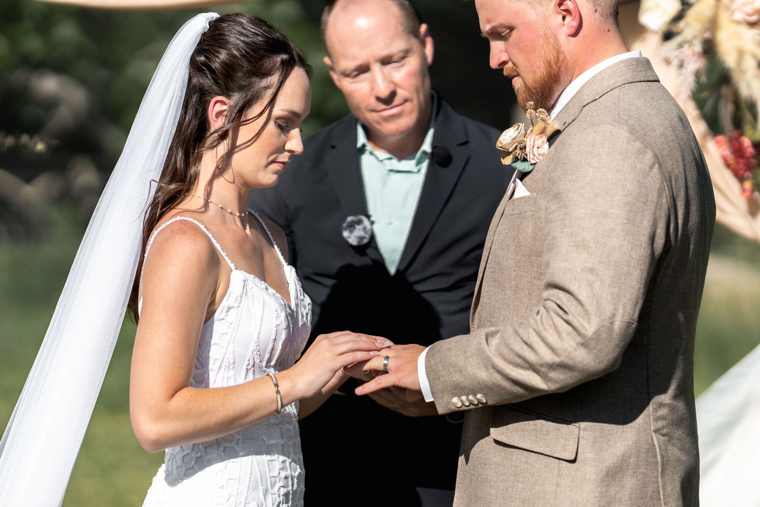 A couple exchanges rings during an outdoor wedding ceremony as the officiant stands between them.