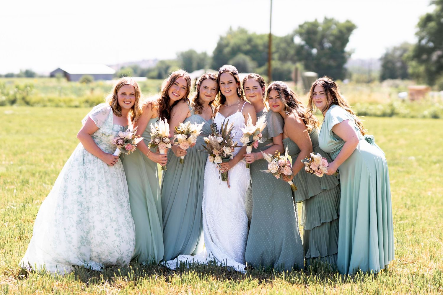 Bridal party wearing pastel blue and mint dresses pose together holding bouquets in a sunny meadow.