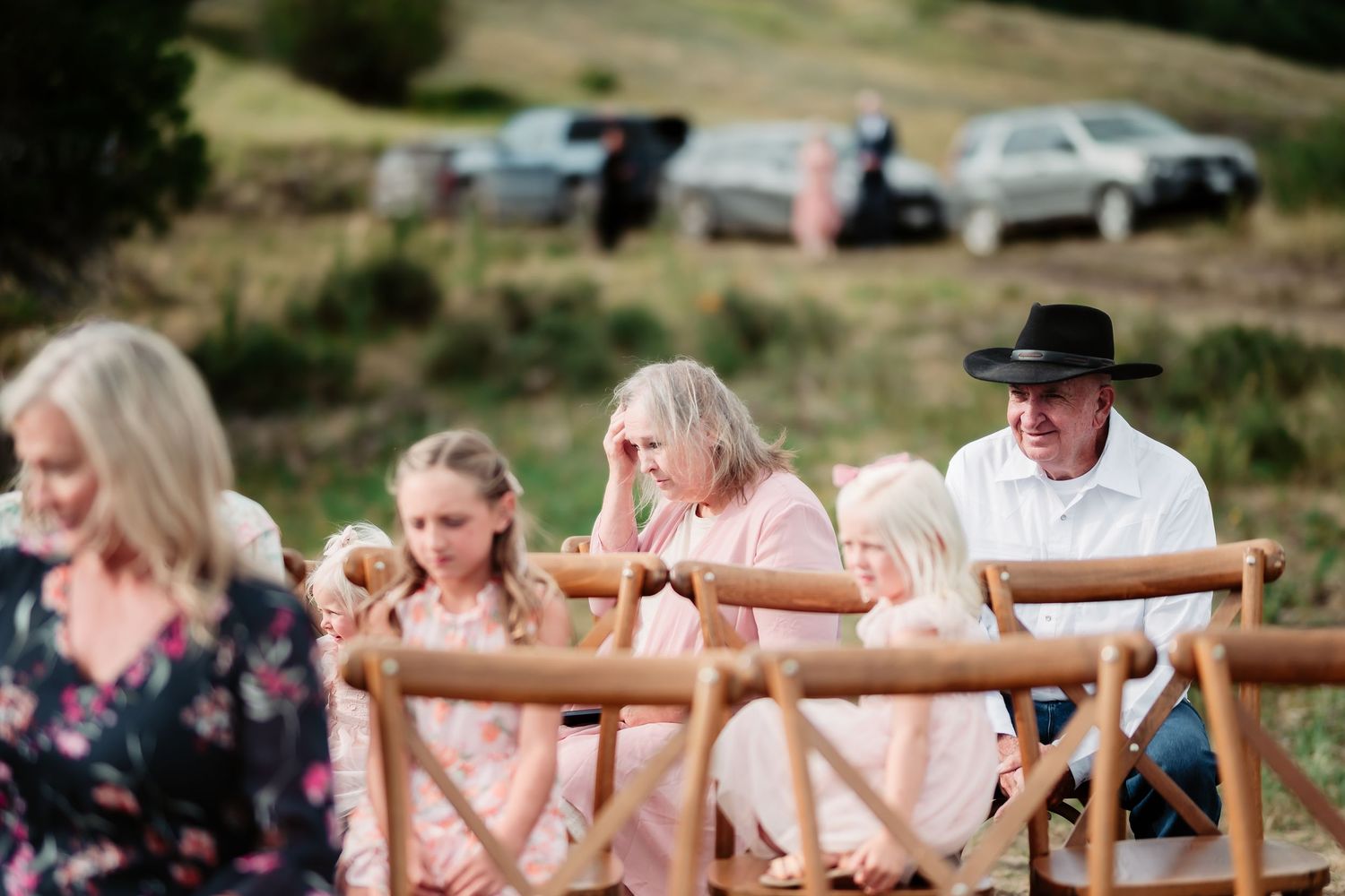 Guests seated in wooden cross-back chairs watching an outdoor wedding ceremony on a hillside.