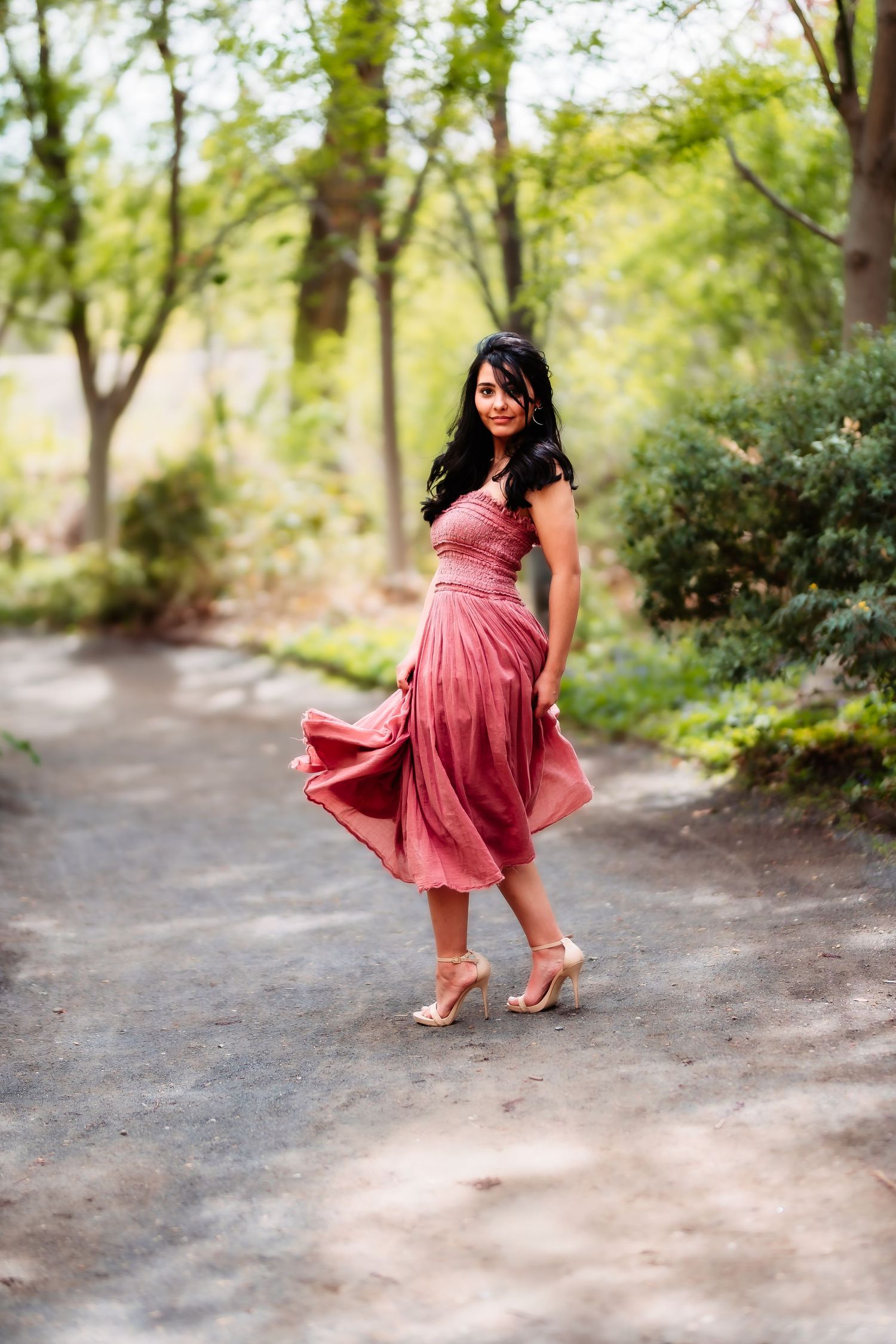 A person in a pink midi dress twirls on a garden path surrounded by green foliage.