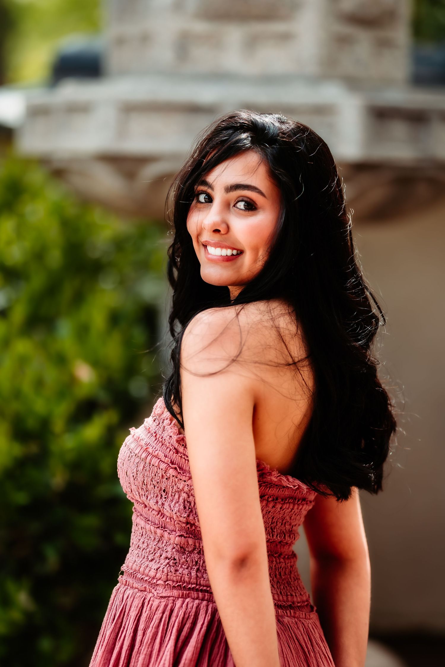 A portrait of someone in a textured pink dress smiling warmly against a blurred stone background.